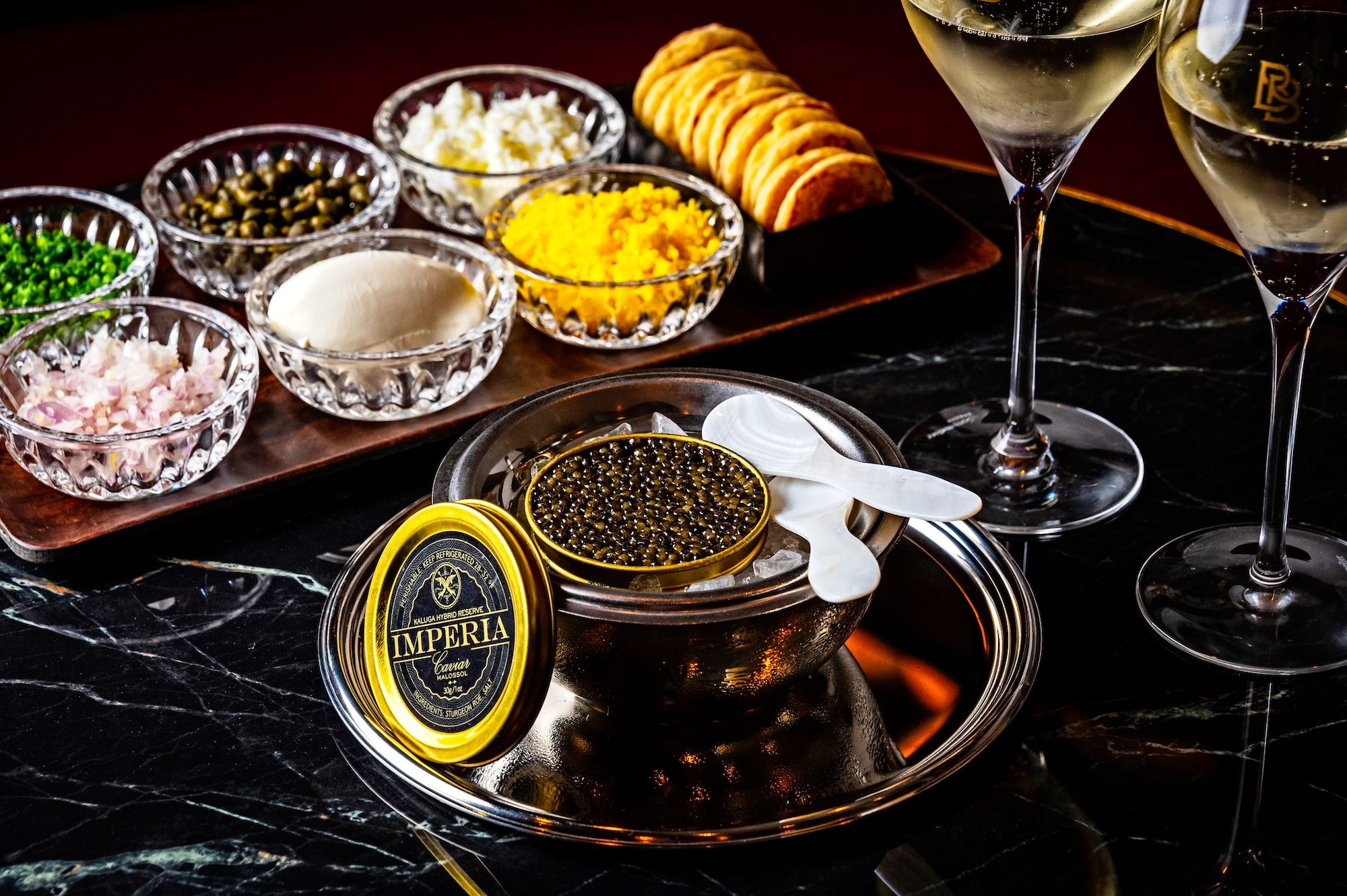 Opened tin of black caviar on ice with white mother-of-pearl spoons, surrounded by small glass bowls of chopped onions, chives, capers, cream, and egg yolk, plus crackers and two glasses of white wine.