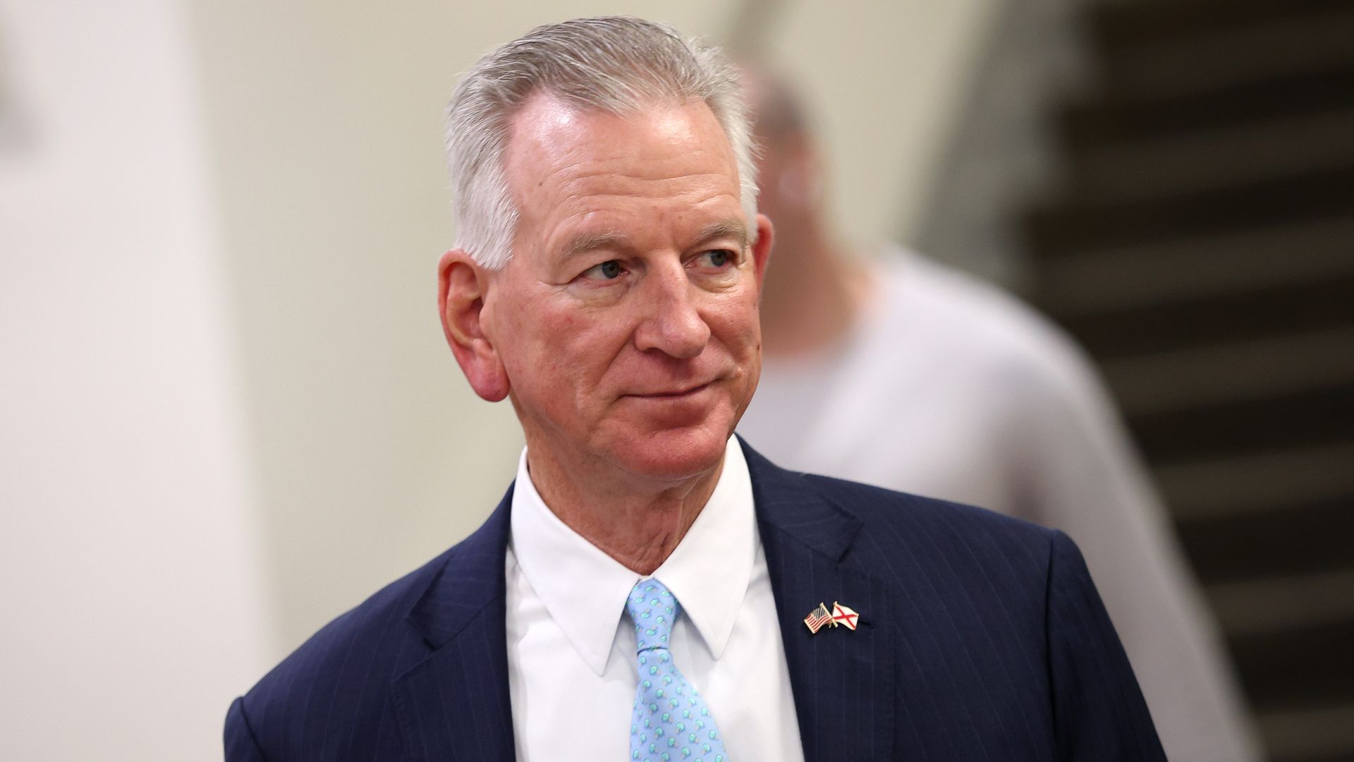 A man with short gray hair in a dark pinstripe suit and light blue tie, looking to the left. He wears two small lapel pins, indoors with a blurred staircase in the background.