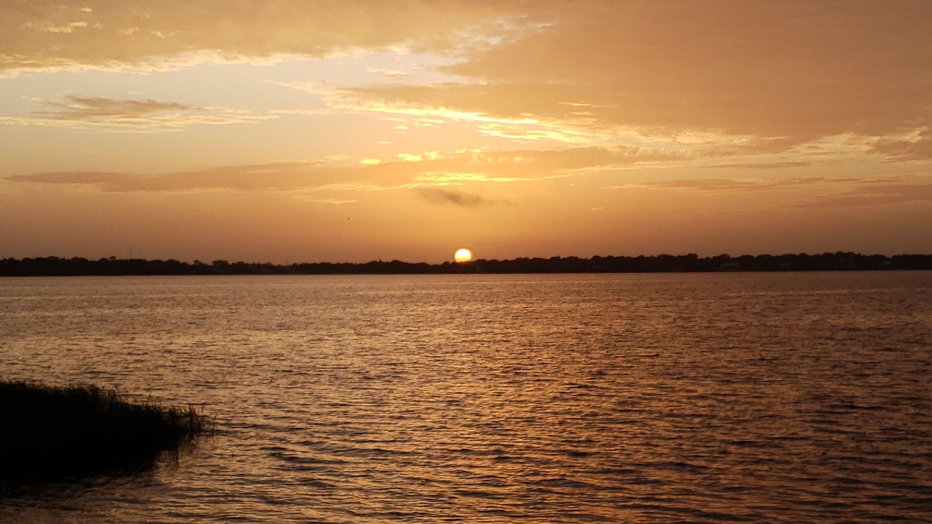 Sunset over a calm body of water with gentle ripples, silhouetted shoreline in the distance, and a sky filled with orange and golden clouds.