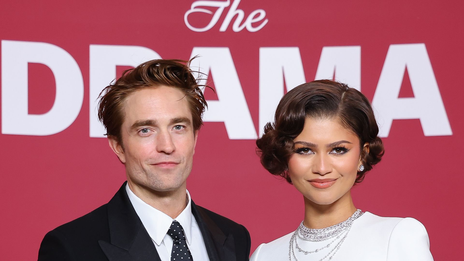 Actor Robert Pattinson wears a black suit and poses arm-in-arm with Zendaya in a longsleeve white dress.