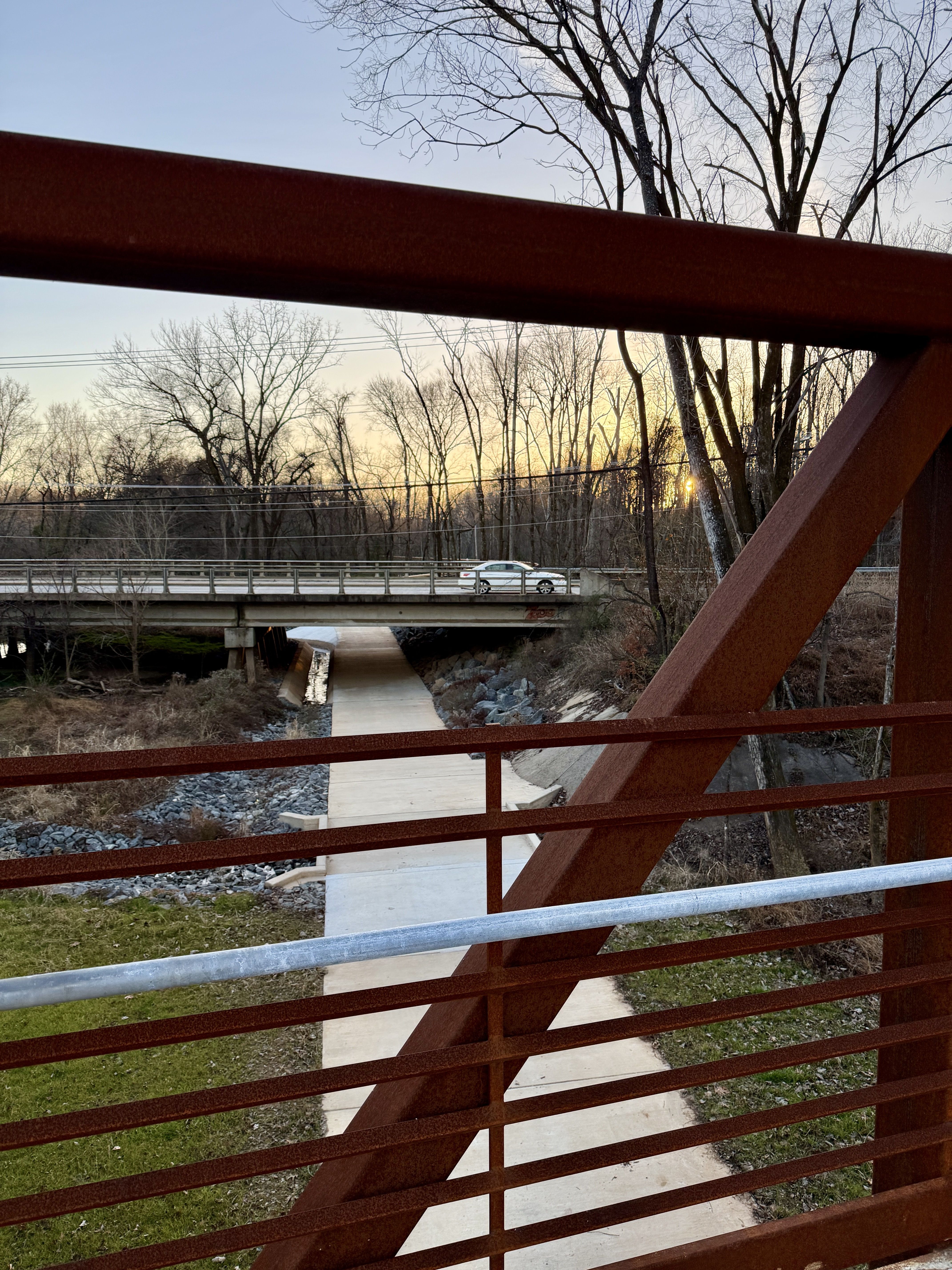 View through rusty red metal bridge rails of a paved path leading to a concrete road bridge with a white car, leafless trees, and a sunset sky in the background.