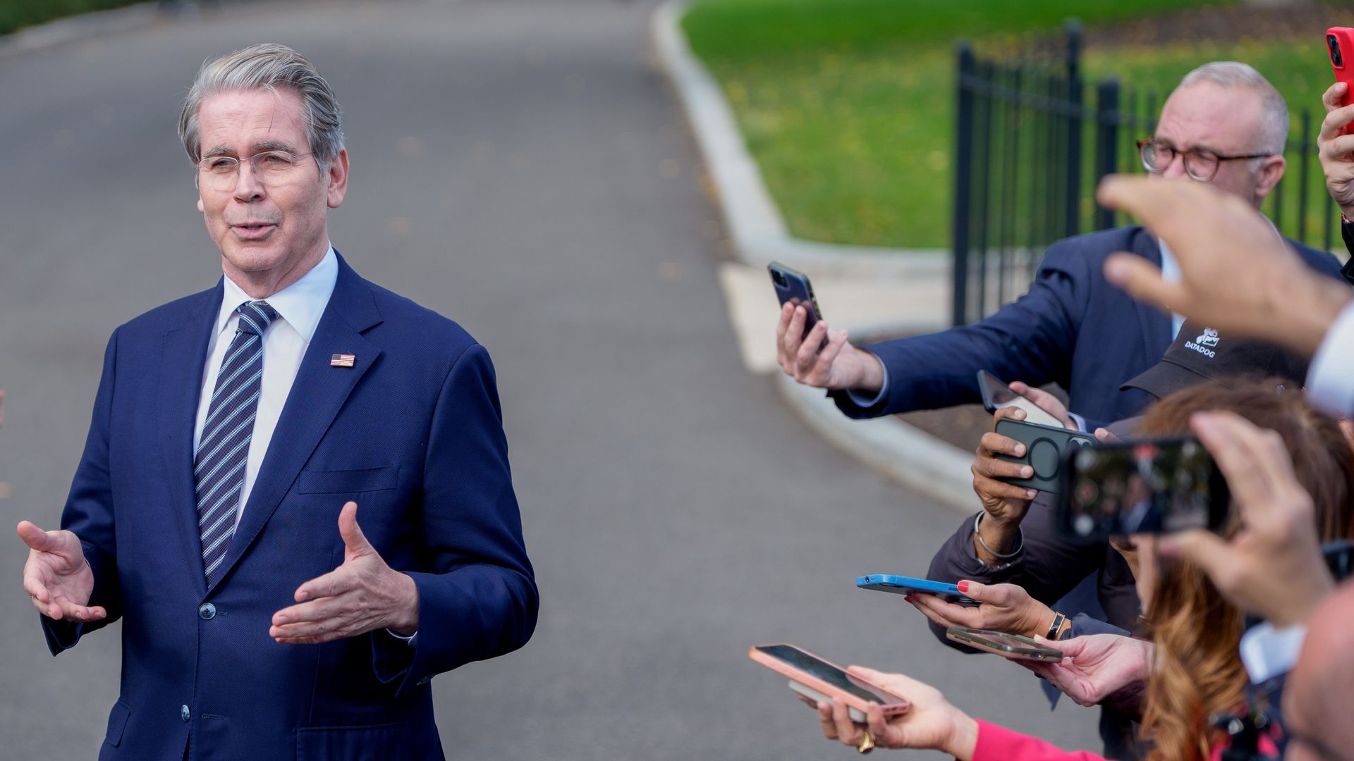 Treasury Secretary Scott Bessent, in a suit and tie, gestures outward with his hands as reporters to his left point their phones at him. 