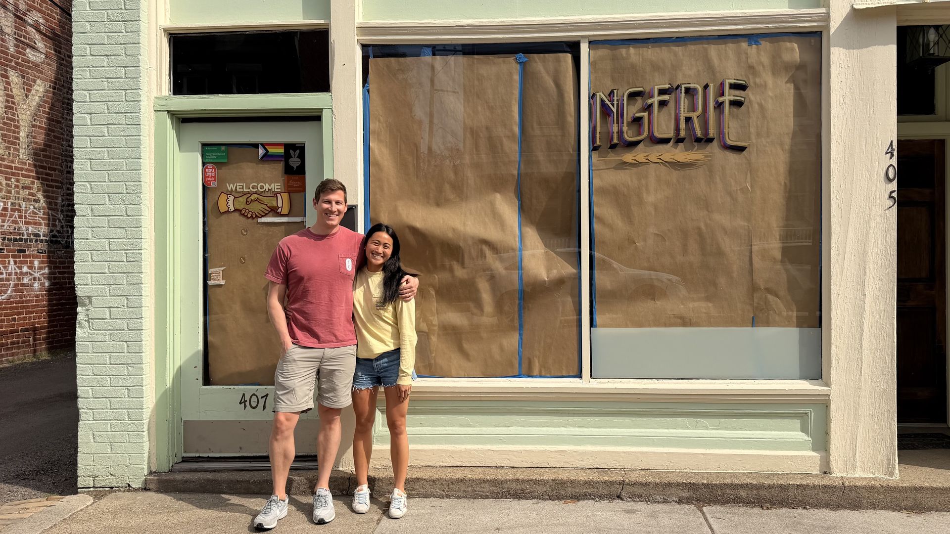 Two smiling people, a man in a red t-shirt and a woman in a yellow top, stand arm-in-arm in front of a pastel storefront with brown paper covering the windows, blue tape, and a faded sign.