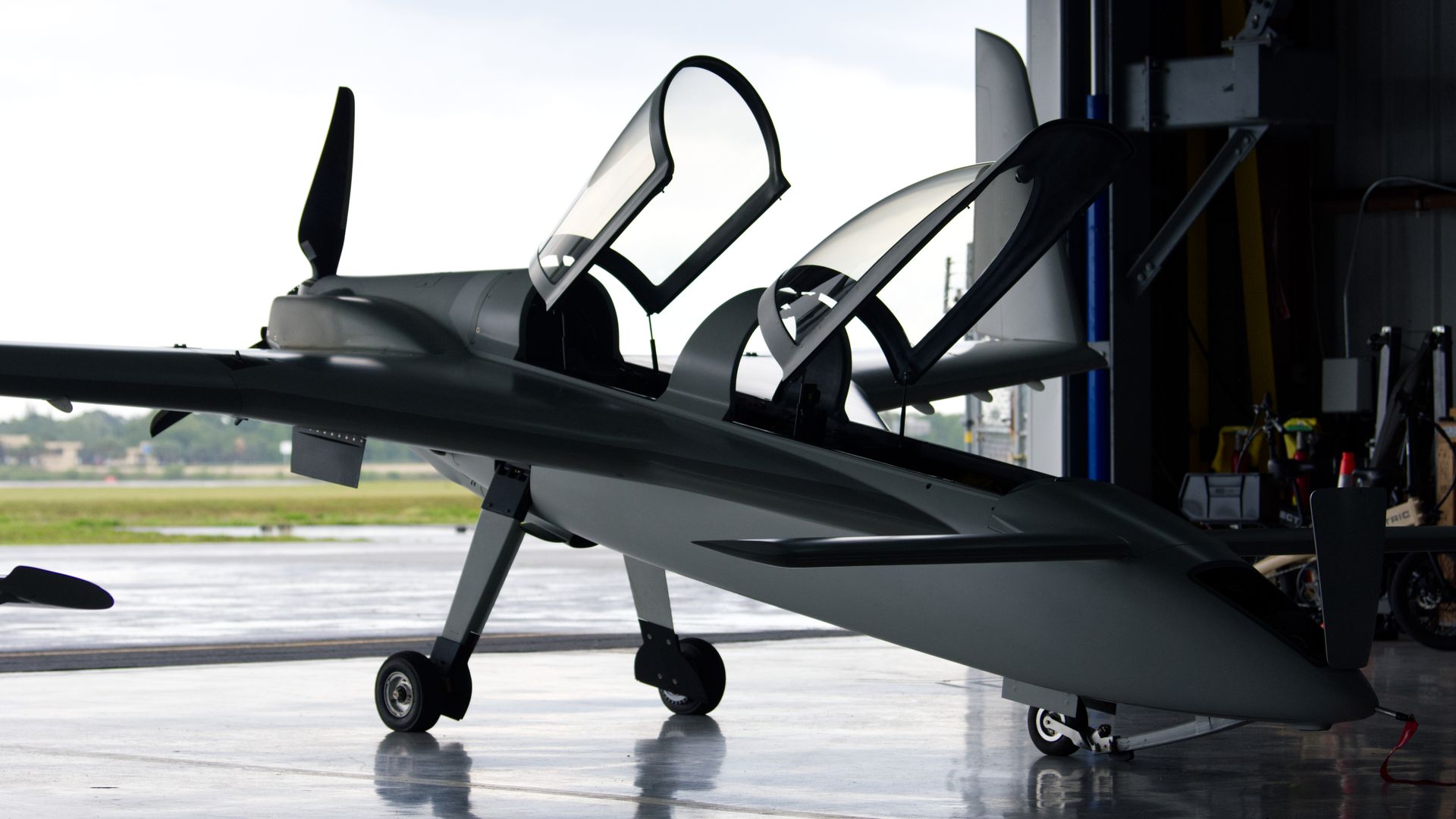 An experimental two-seater aircraft sits in a dimly lit hangar. There is a large, open door in the background, and rain can be seen outside.