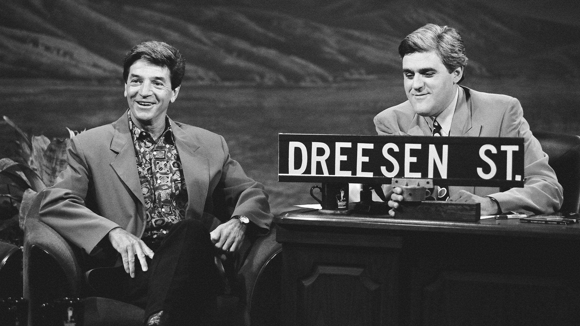 Black-and-white photo of two men in suits on a talk-show set, smiling; one holds a sign reading "DREESEN ST" beside a desk.