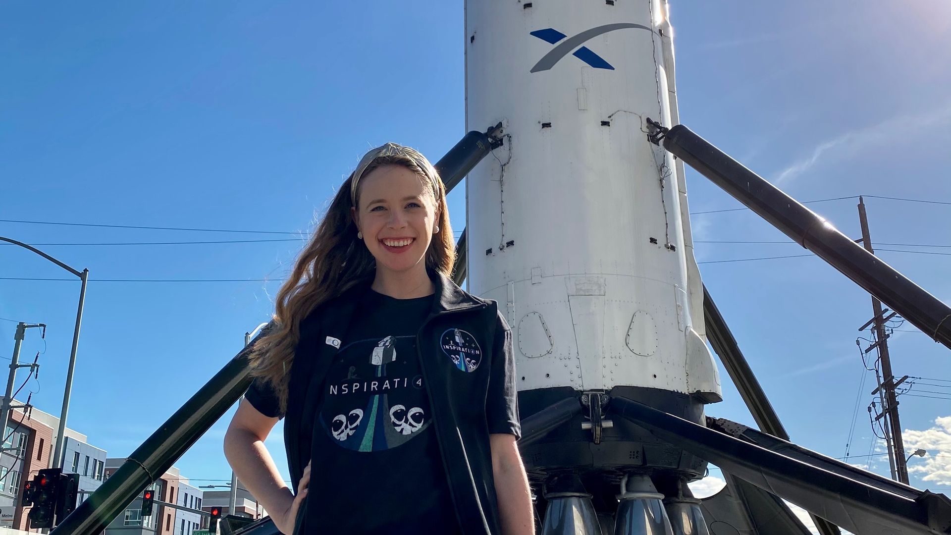 A white woman stands in front of a SpaceX rocket with her hand on her hip, smiling.