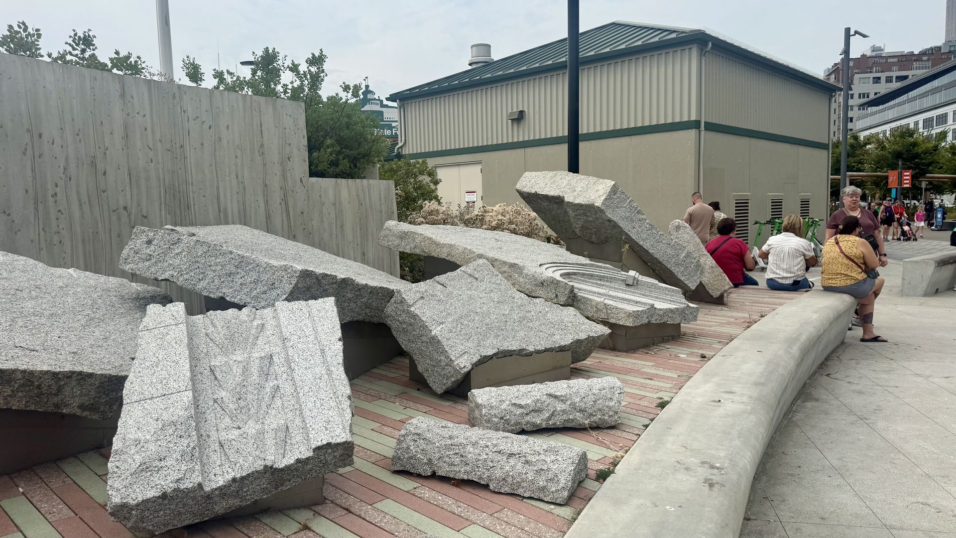 An outdoor urban area with large jagged pieces of rock shaped like architectural elements arranged along an elevated brick platform. In the background is a beige industrial building with green trim; people sit and stroll nearby.