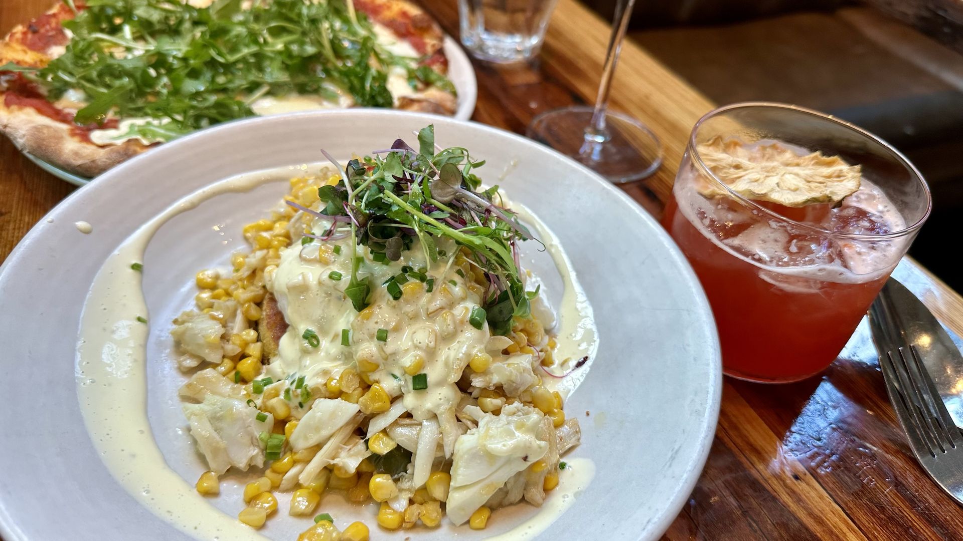 Eggs benedict with corn kernels and microgreens, served on a gray plate. Background shows pizza with arugula and a red cocktail with a dried pineapple slice on a wooden table.