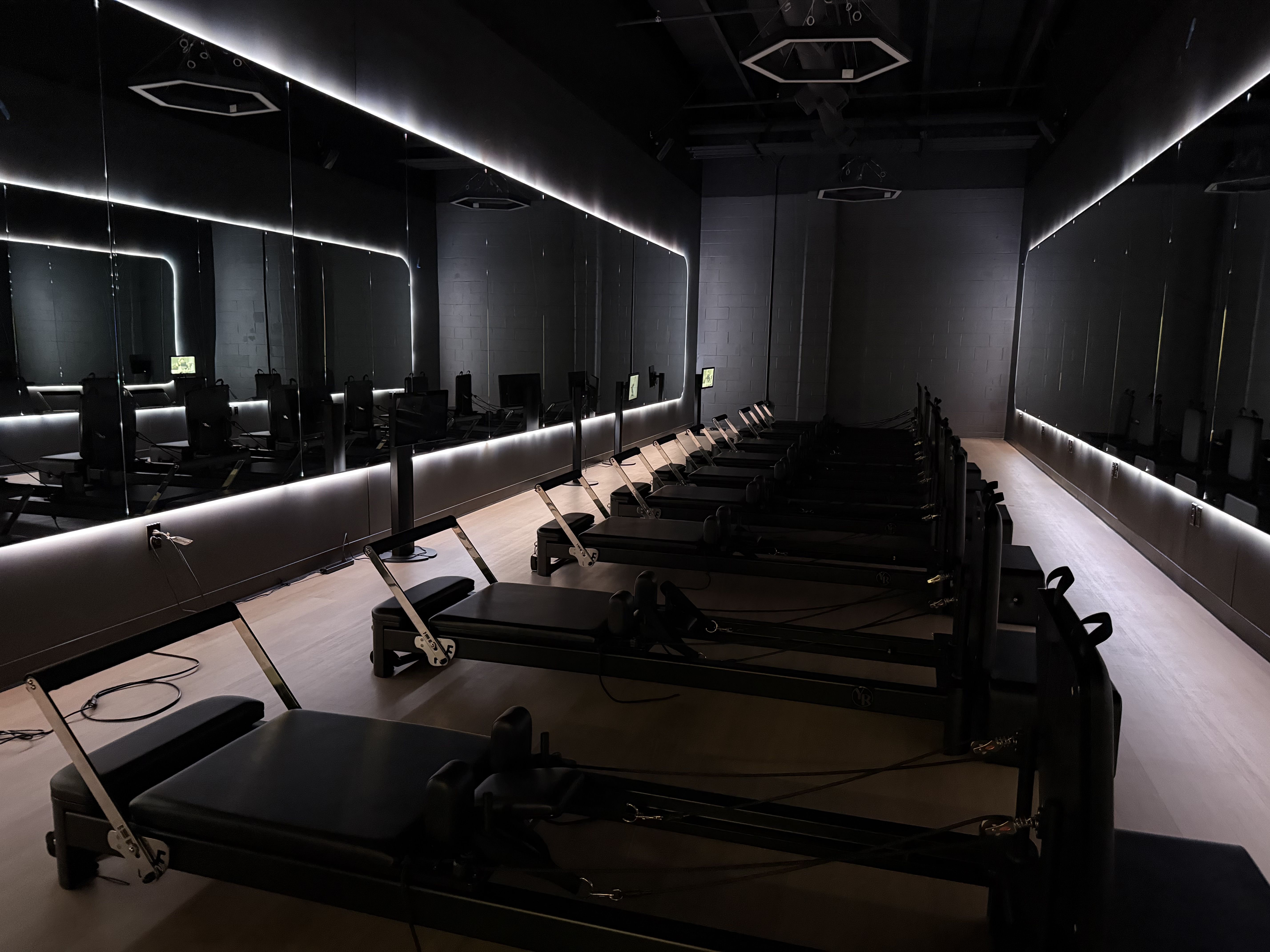 Dimly lit Pilates studio with rows of reformer machines along mirrored walls, glowing white LED edge lights, and a wooden floor; dark ceiling with geometric fixtures.
