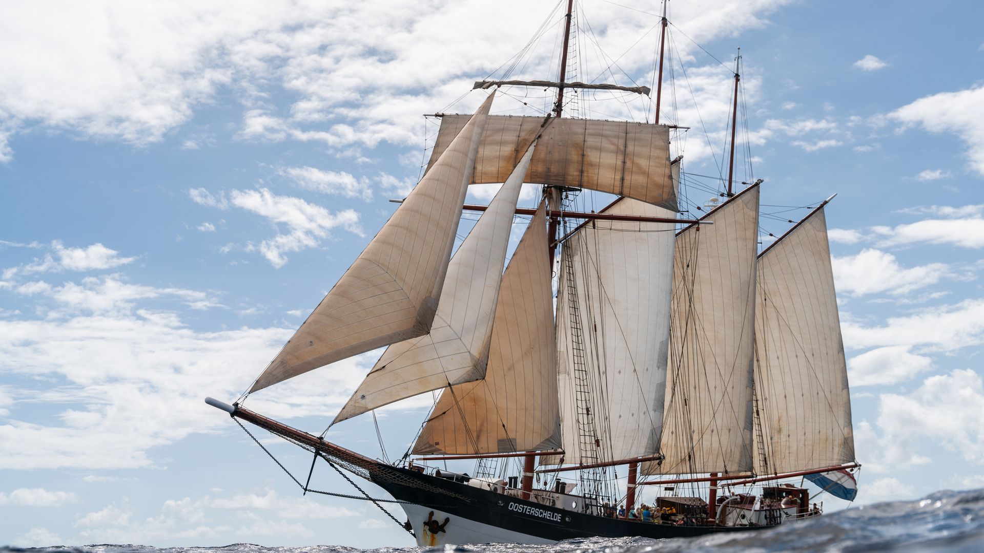 Tall sailing ship with cream canvas sails, brown masts and complex rigging, gliding on a blue ocean under a partly cloudy sky; crew visible on the deck.