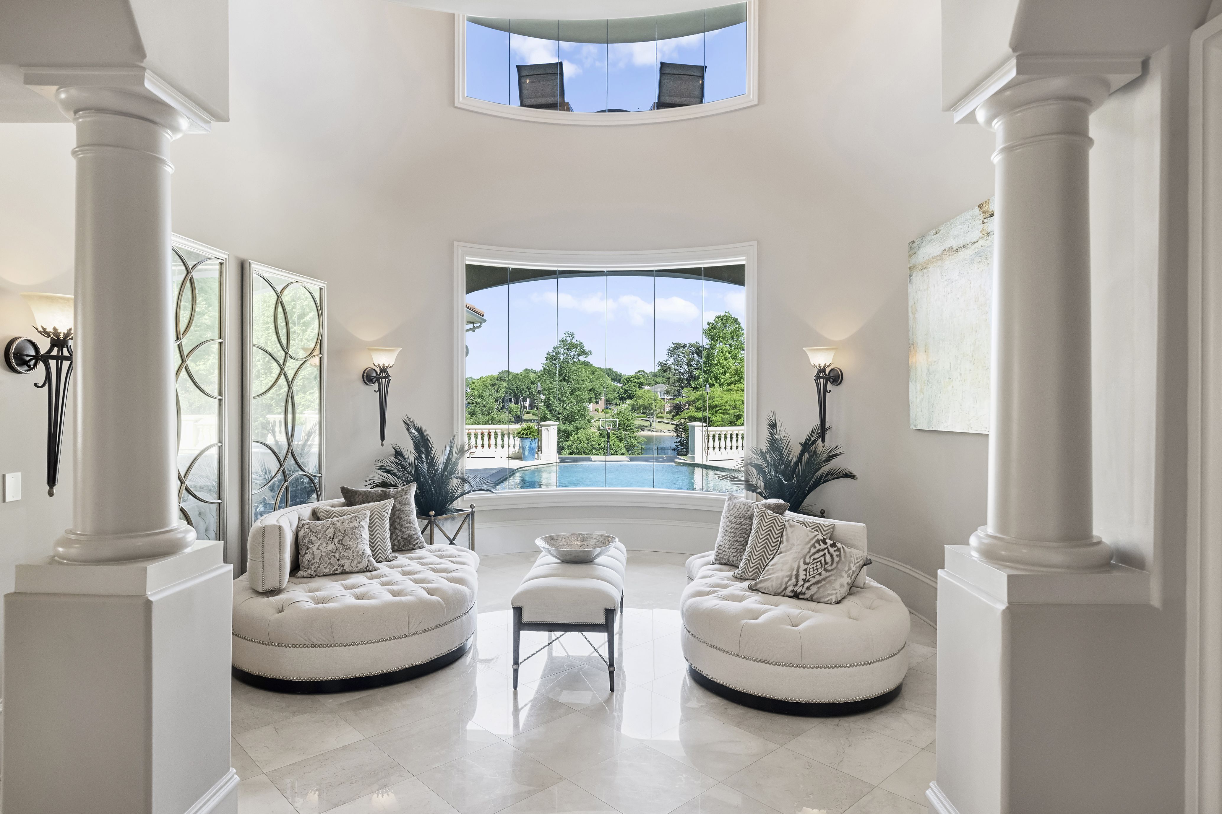 Luxurious living room with two large white columns flanking a curved bay window, overlooking a pool and trees; tufted white seating, decorative pillows, and wall sconces create an airy, elegant space.