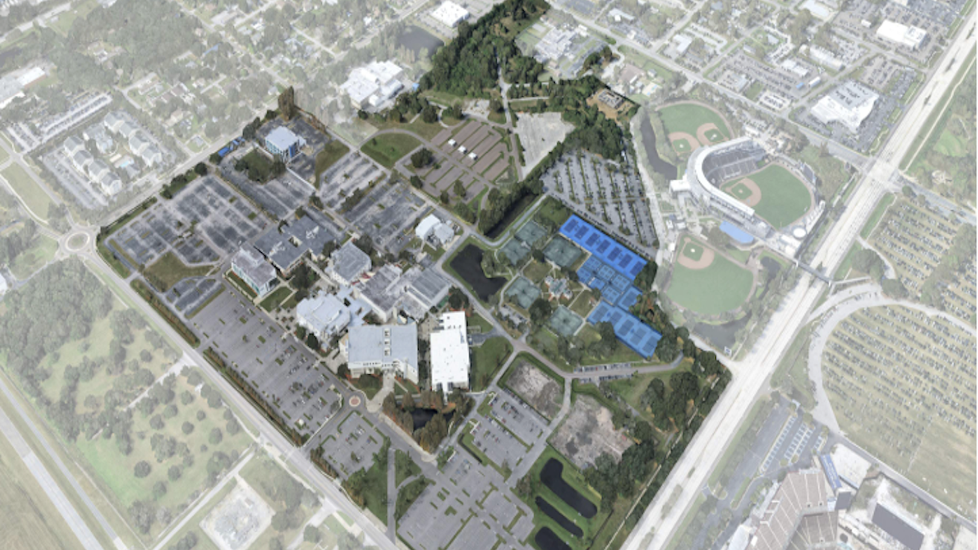 Aerial view of a large complex with parking lots, multiple buildings, blue tennis courts, two baseball fields, green spaces, and surrounding roads in a suburban area.