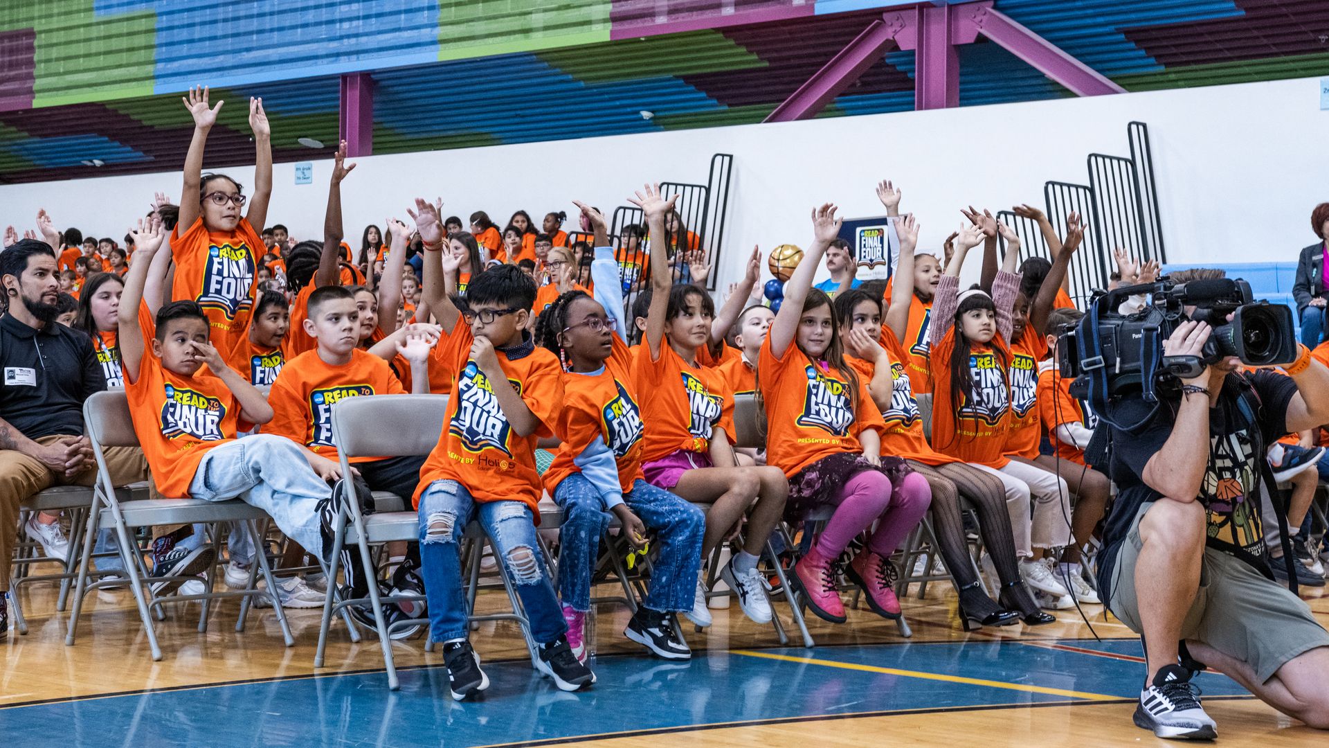 Group of children in bright orange "Read to the Final Four" shirts sitting on chairs in a gym, many raising their hands while a cameraman films on the right side.