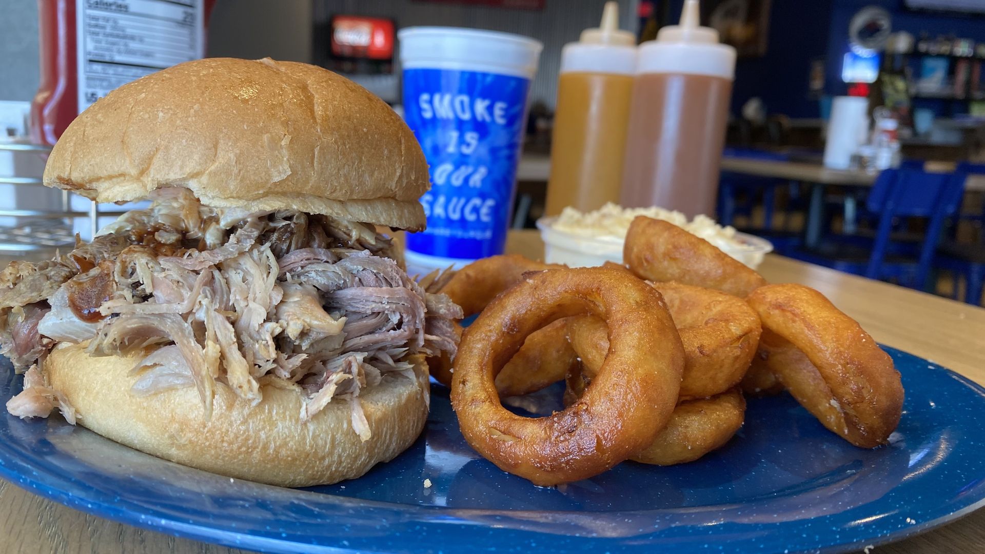 A photo of a bbq pork sandwich and onion rings on a blue plate with sauces and a drink in the background.
