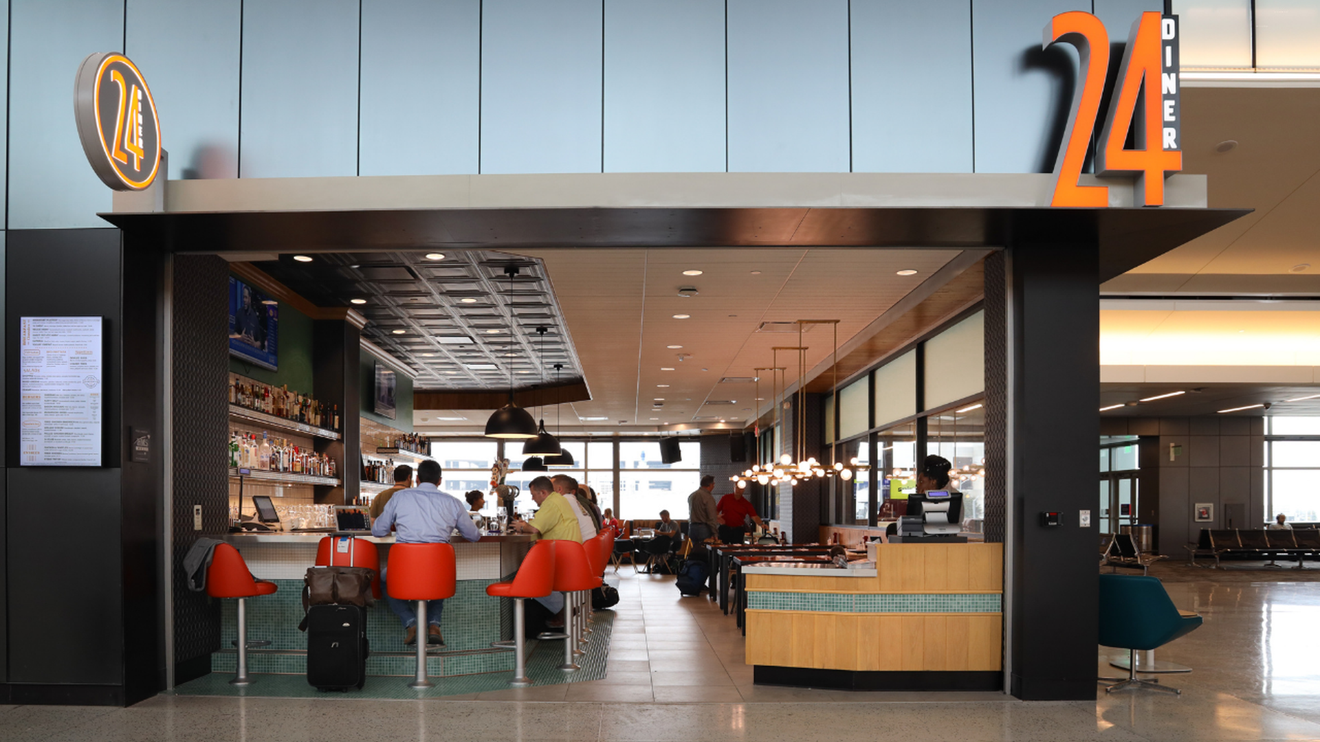Interior of 24 Diner with orange bar stools and patrons seated at a bar and tables, modern lighting, and a large orange "24" sign above the entrance in a bright public space.