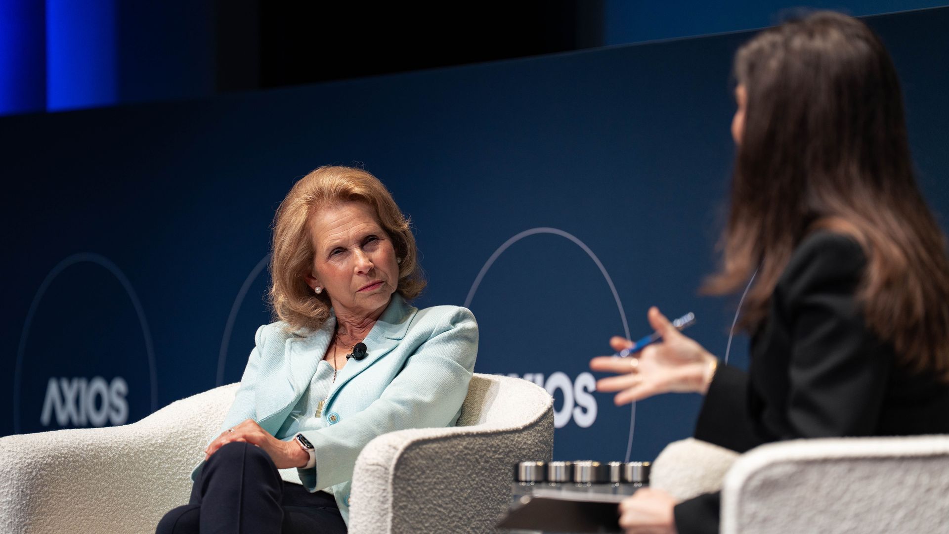 Two women sit in white armchairs on a stage with a dark blue background featuring the word "AXIOS." Shari Redstone in a light blue blazer listens intently to Axios' Sara Fischer in black.