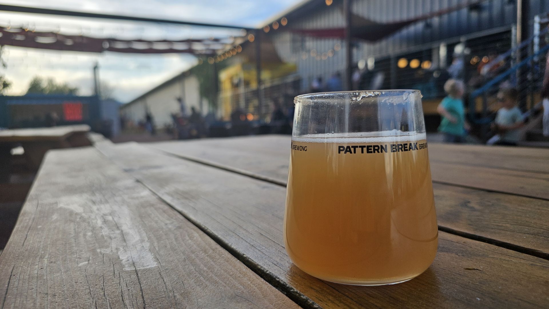 A pint of hazy beer on a wooden table at a brewery patio; string lights and blurred people in the background, with the words "PATTERN BREAK" on the glass.