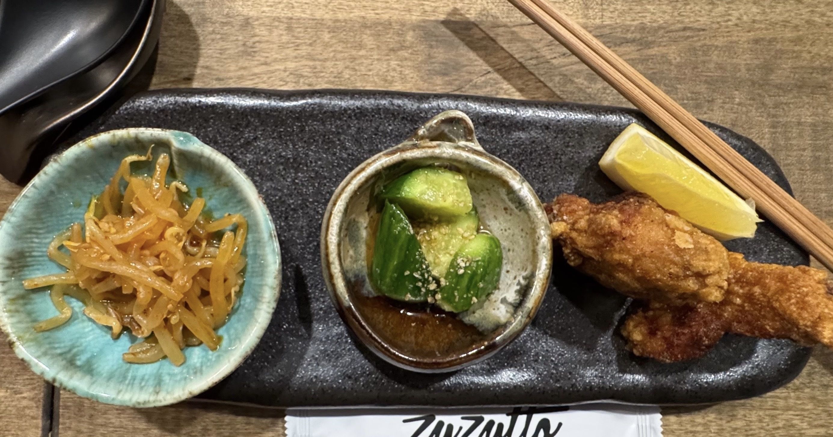 A black platter with small Japanese-style sides: bean sprouts, marinated cucumbers, two fried chicken pieces, lemon wedge, and chopsticks.
