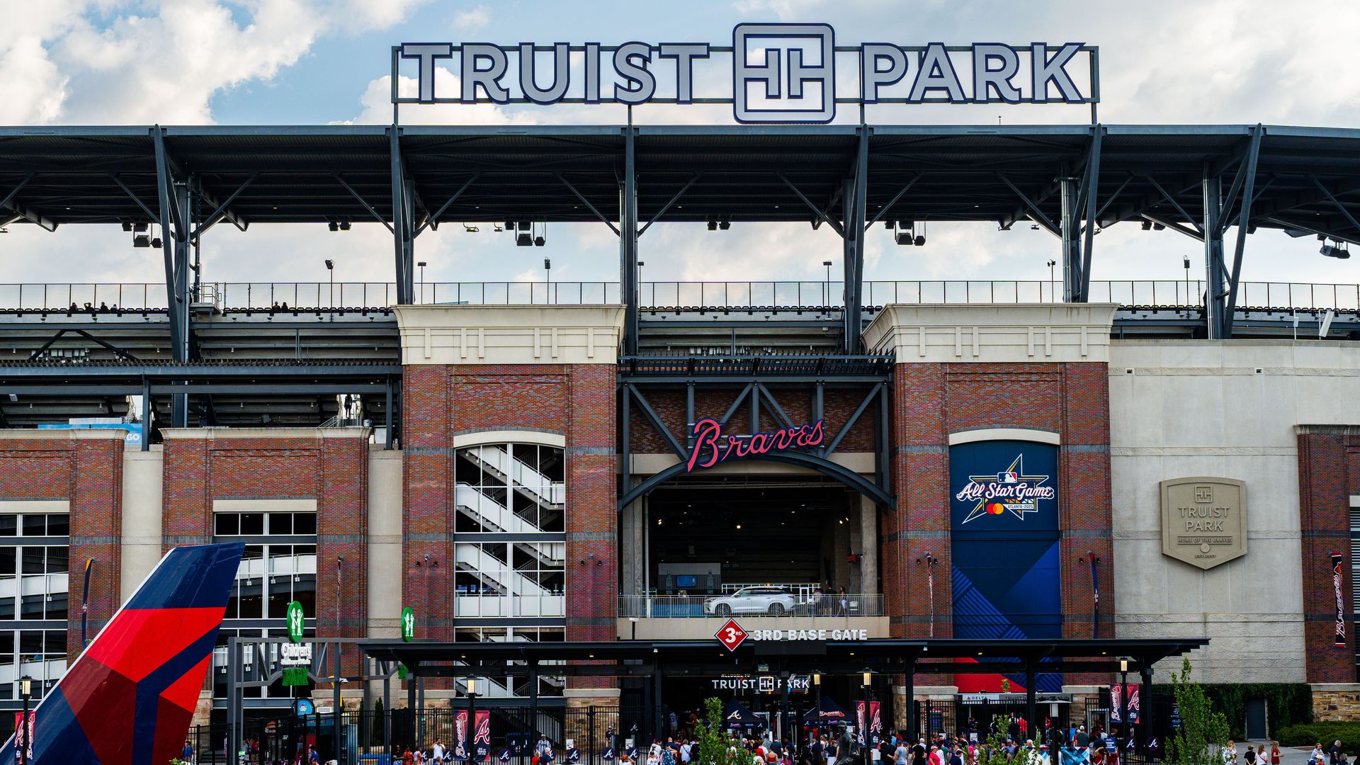 A photo showing the outside of a stadium with a brick exterior. A large sign above the building reads: Truist Park. People are walking in front of the building. The sky is blue with several white clouds.