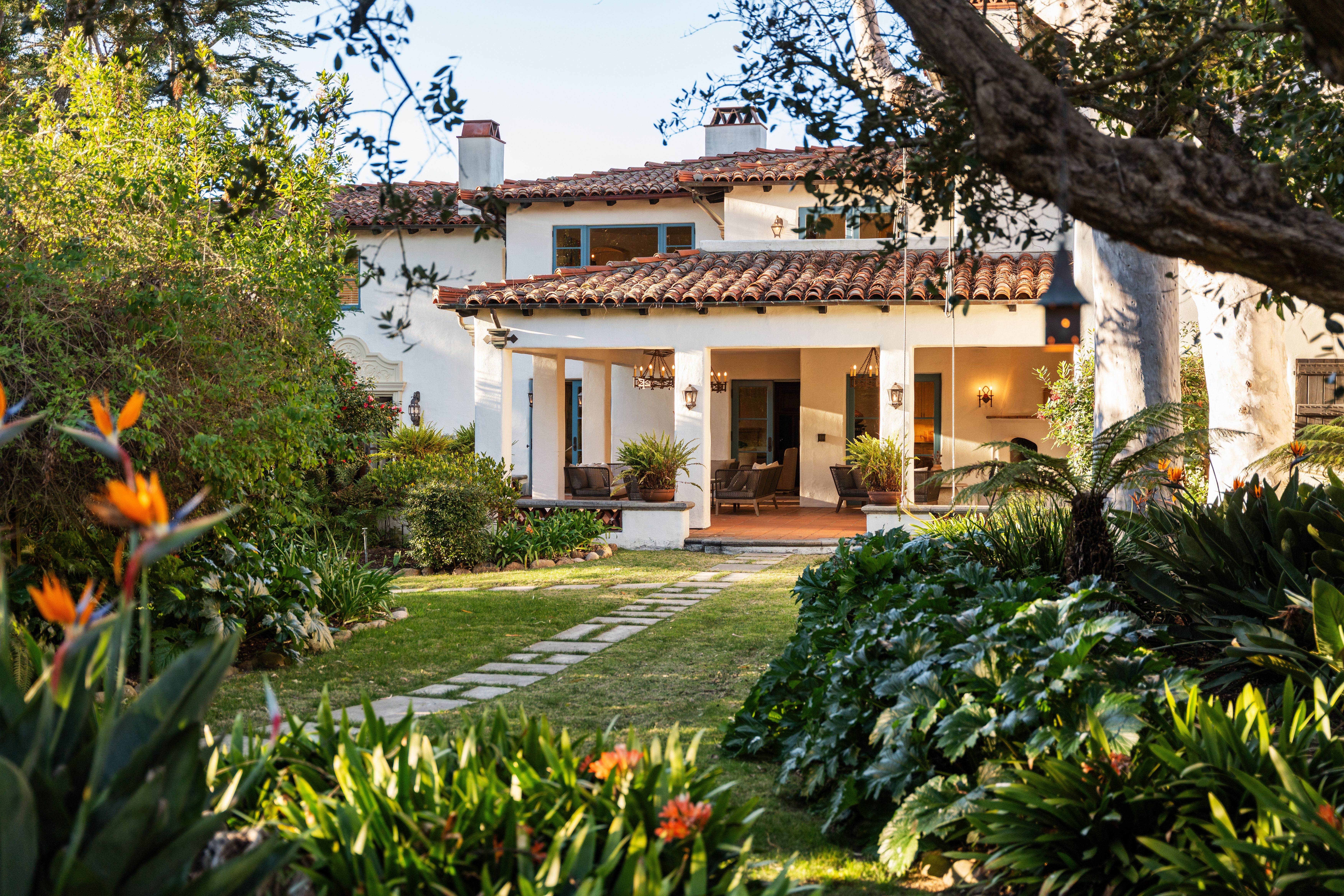 Sunny yard with a white stucco house, red-tiled roof, blue-trim windows, and a covered patio. Lush garden frames the scene with a large tree branch in the foreground.