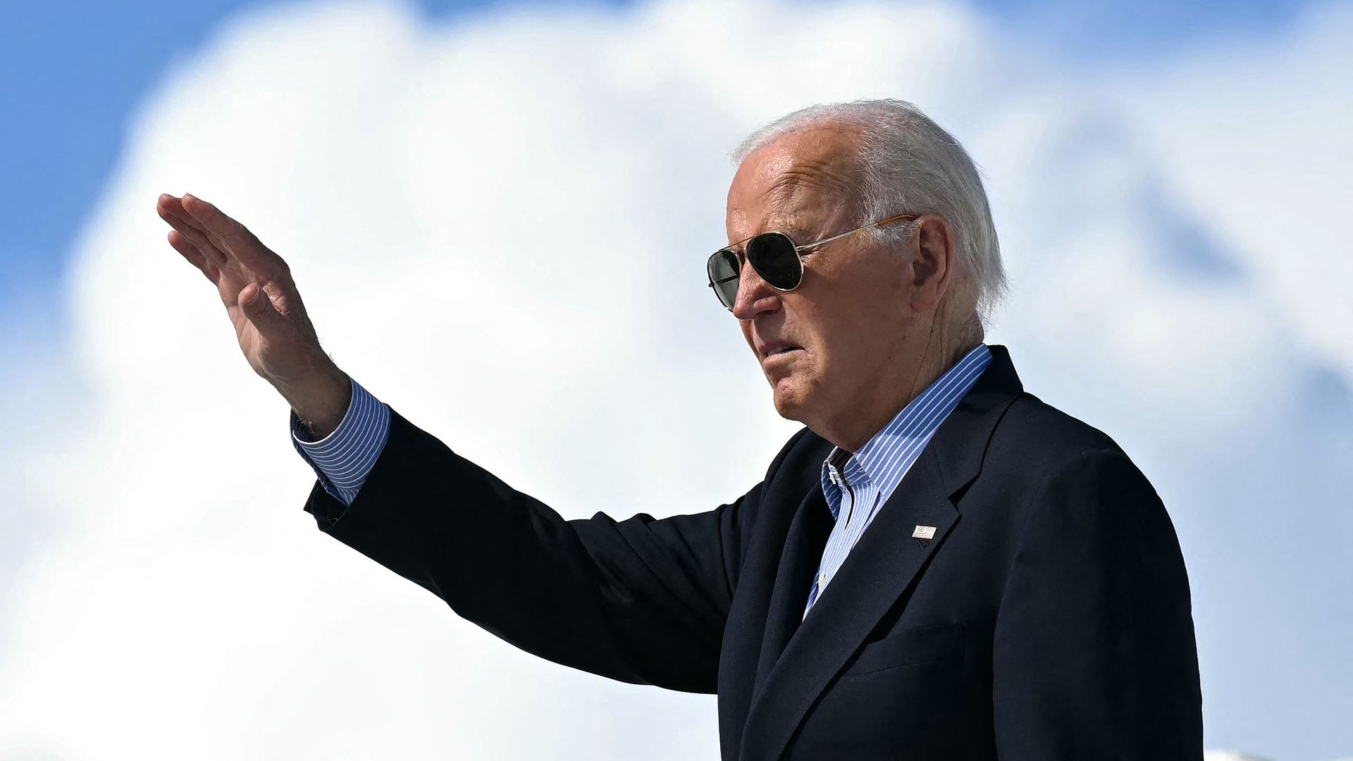 President Biden, waving while wearing a dark gray suit under a blue, cloudy sky.