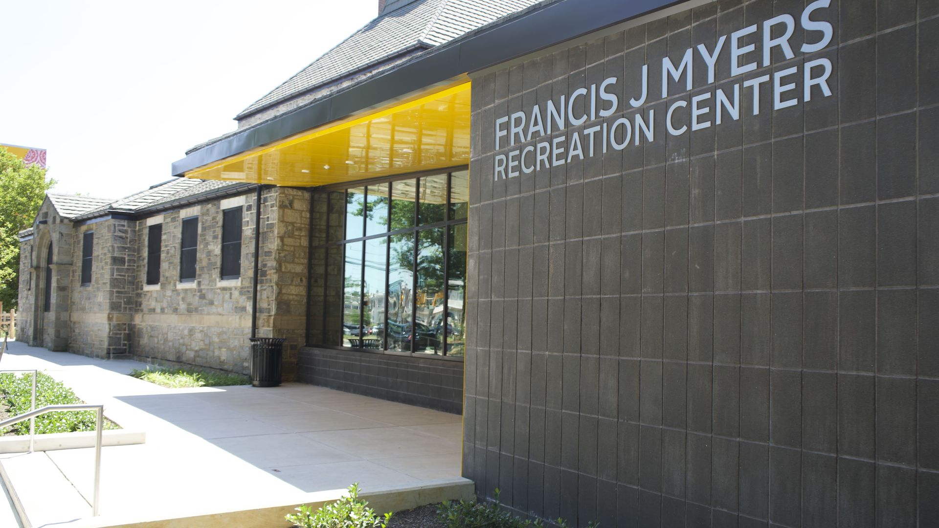 Exterior of Francis J. Myers Recreation Center: dark tiled wall with large white letters, bright yellow canopy, stone facade, glass windows, sidewalk and greenery on a sunny day.