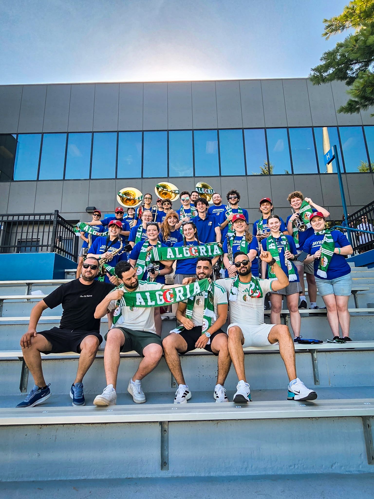Group of Algeria fans in blue shirts and green scarves pose on stadium steps in front of a gray modern building with blue windows, holding ALGERIA banners and brass instruments on a sunny day.