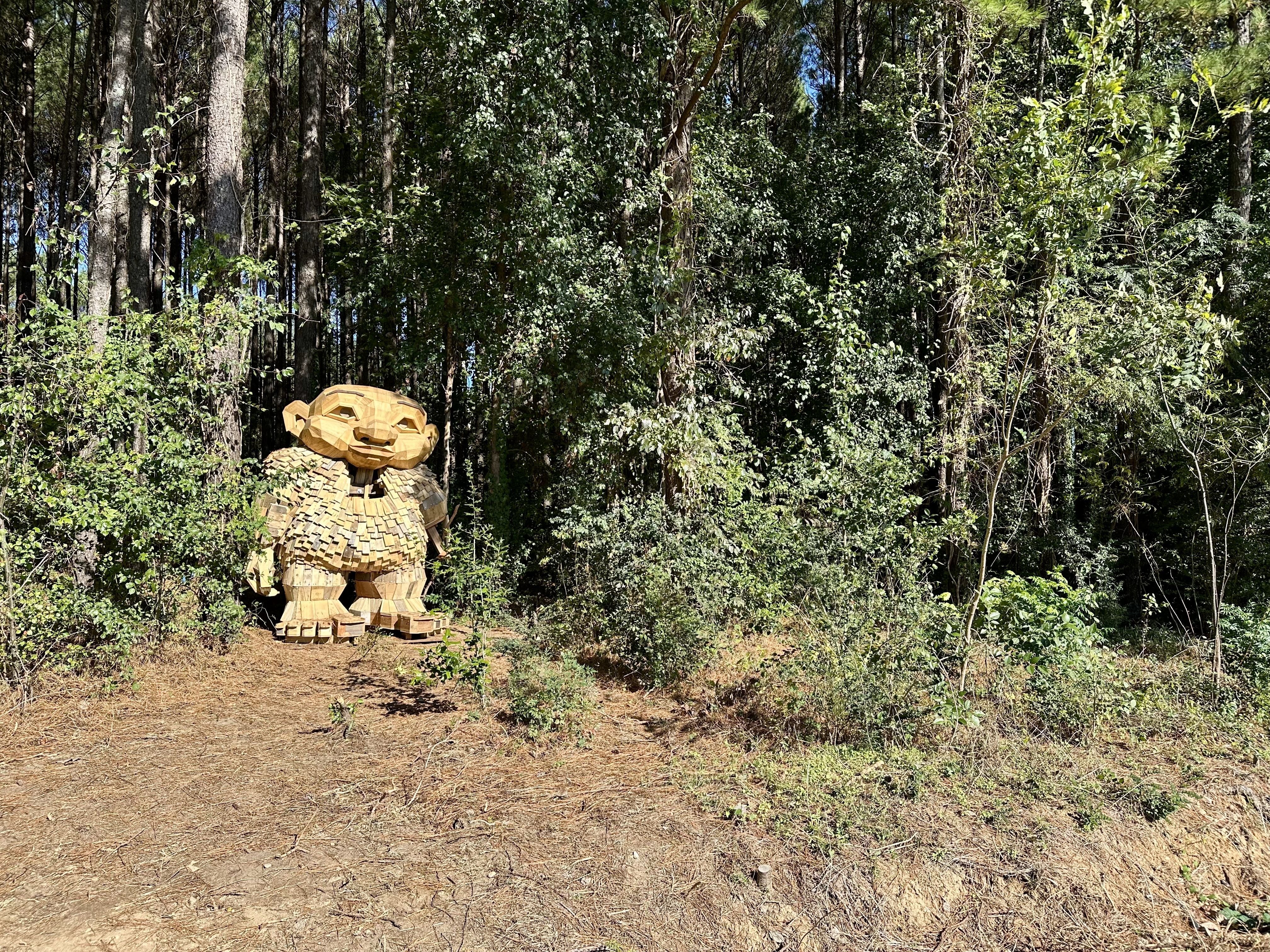Large wooden sculpture of a humanoid figure with a textured robe, standing at the edge of a forest clearing with dense green trees and underbrush around it.