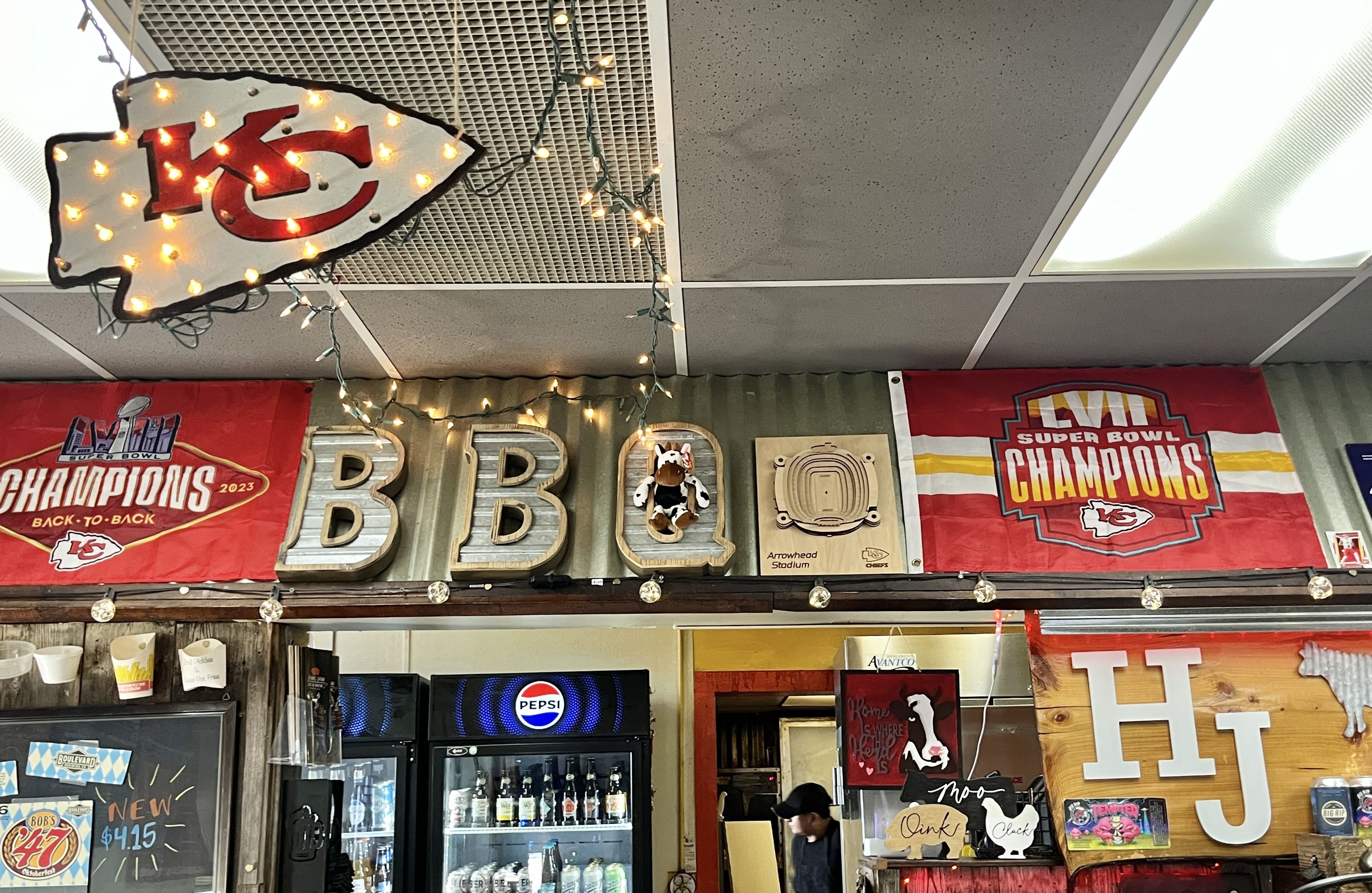 Interior wall with Kansas City Chiefs Super Bowl champions banners, illuminated KC arrow sign, BBQ letters with a cow plush, string lights, Pepsi fridge, and wooden letters HJ.