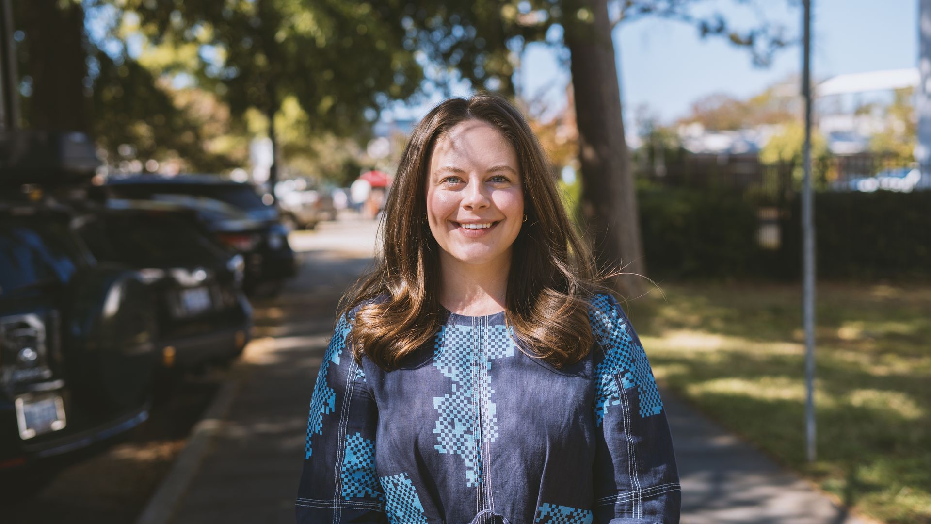 Smiling woman with long brown hair wearing a blue patterned dress, standing on a sunlit sidewalk with trees and parked cars in the background on a clear day.