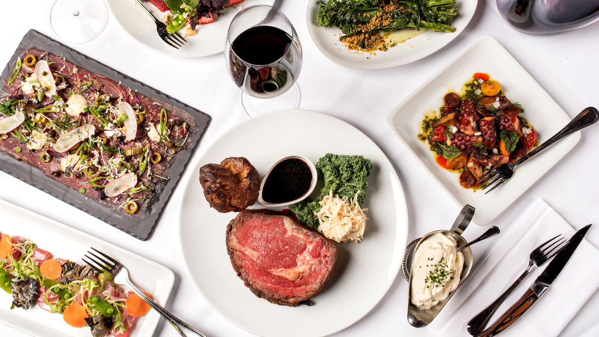 Top view of a gourmet meal with rare steak, Yorkshire pudding, kale, sauce, grilled broccolini, octopus with vegetables, mixed greens salad, and two glasses of red and white wine on a white tablecloth.