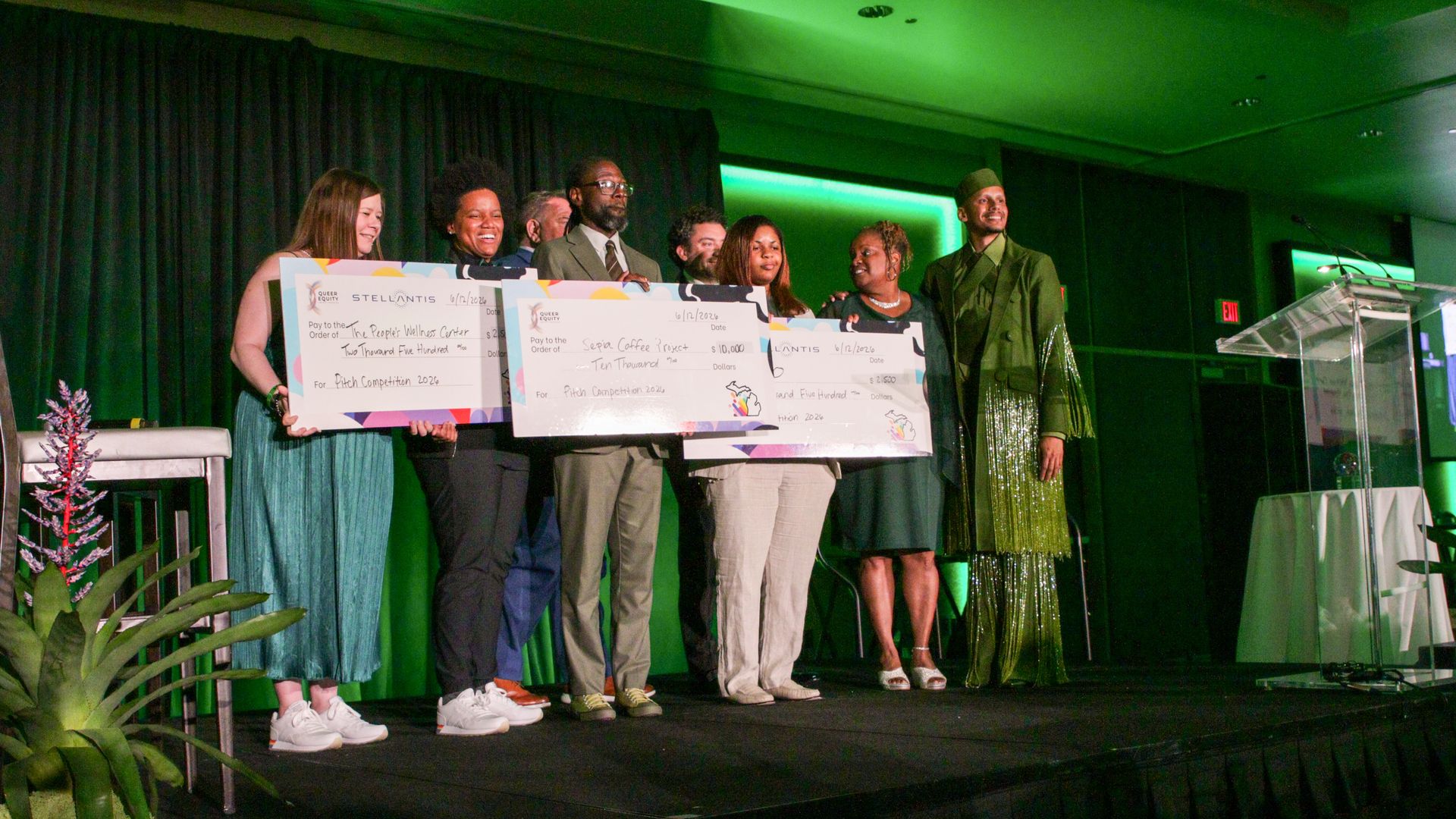 Group of people on a stage holding oversized ceremonial checks, smiling; green stage lighting, dark curtains, and a clear podium on the right.