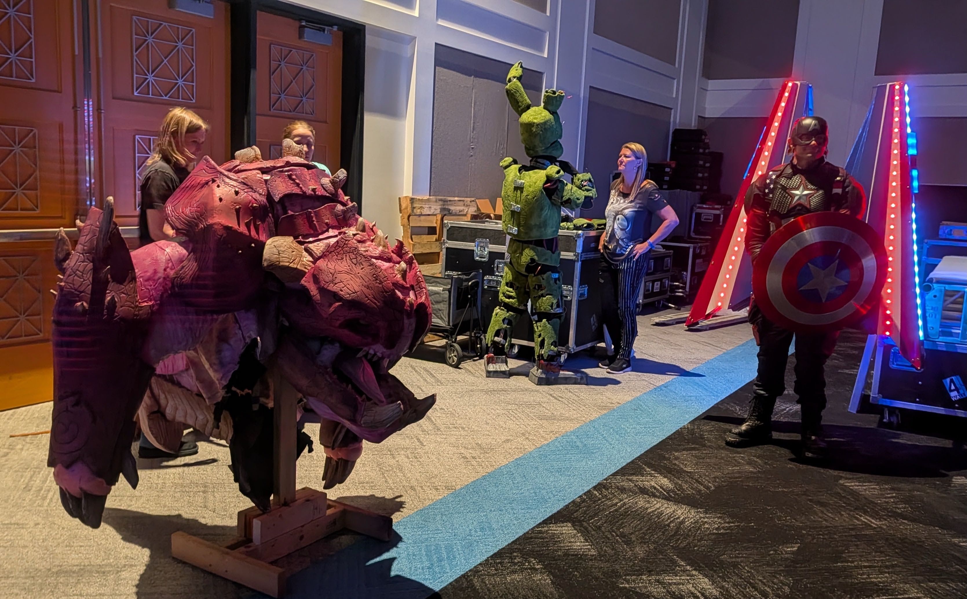 Backstage scene with cosplayers: a large pink rock monster on the left, a tall green alien in the center, and Captain America with shield on the right; lit set pieces in the background.