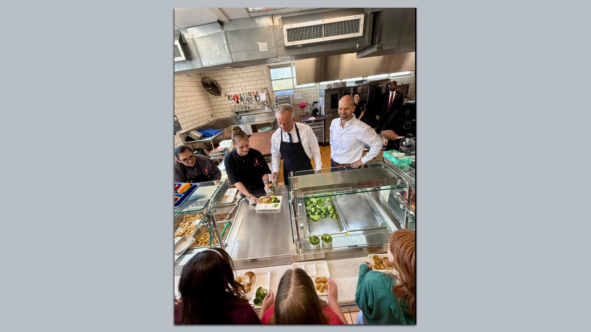 Volunteers serve food including green vegetables and fried items to people in a cafeteria-style kitchen with stainless steel counters and white tiled walls.