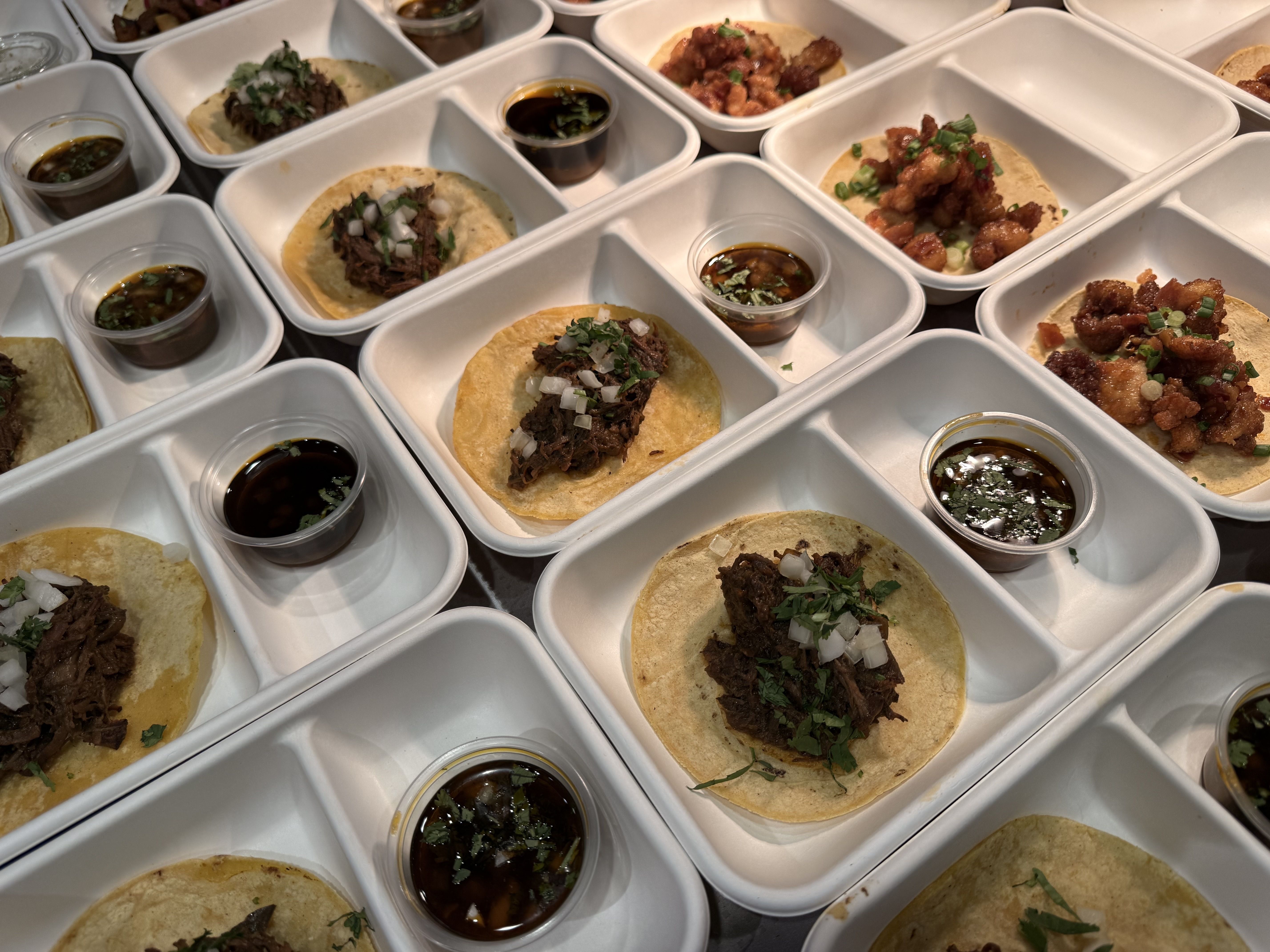 Rows of white takeaway boxes hold soft corn tortillas topped with shredded beef, onions, and cilantro, plus small cups of dark dipping sauce.