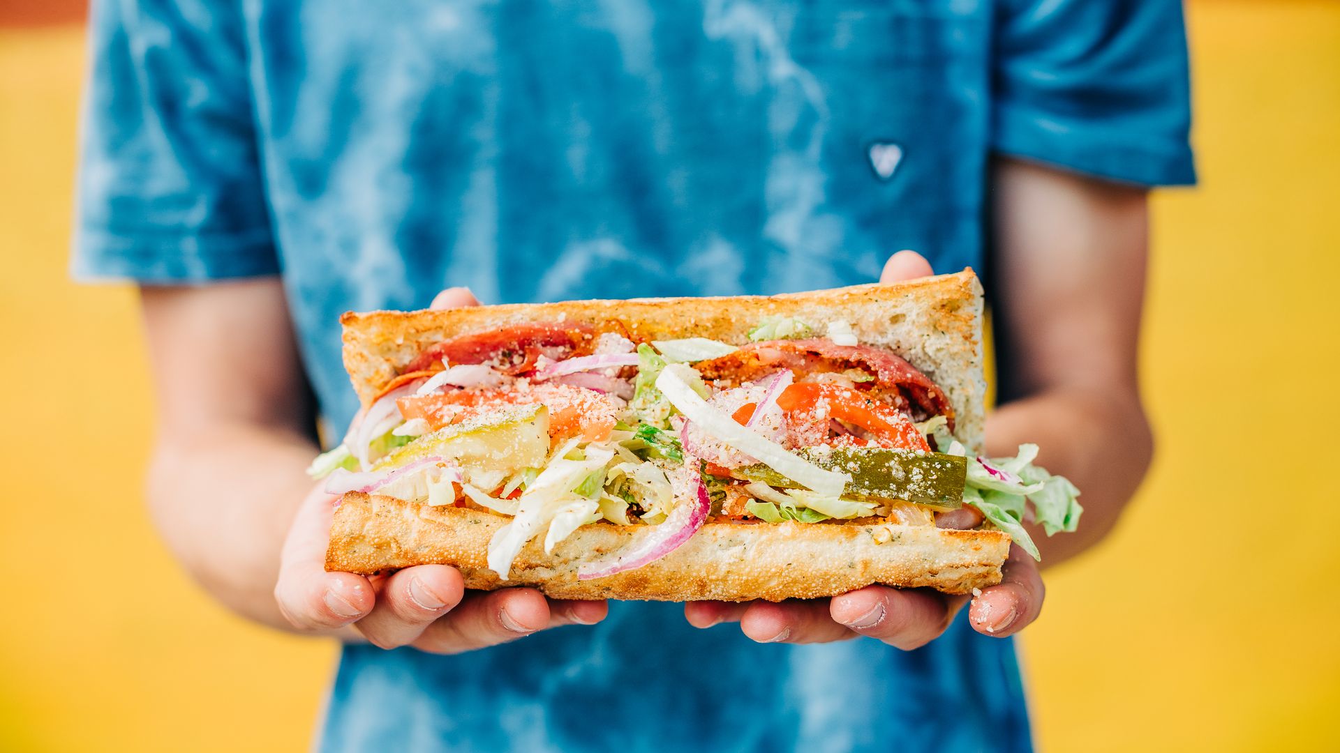 Person in a blue tie-dye shirt holds a large veggie sandwich filled with lettuce, tomatoes, onions, and pickles, against a bright yellow background.