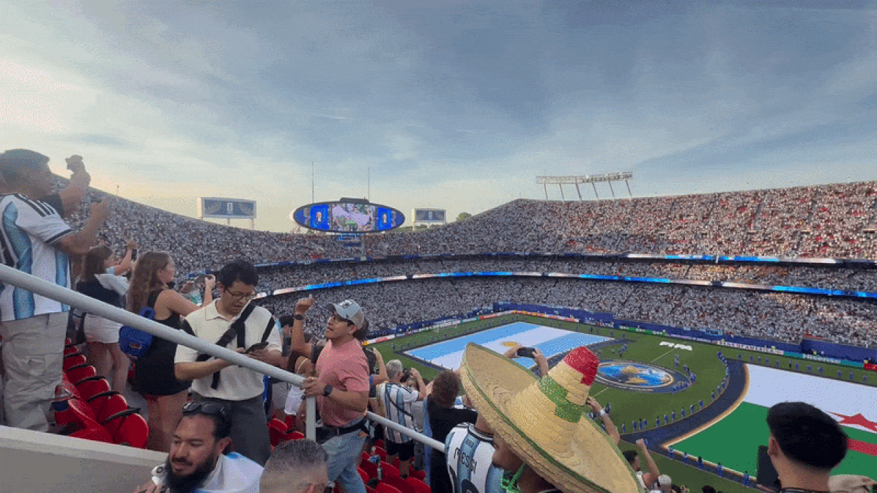 Panoramic view of a packed stadium with a giant center screen and blue-white field graphics; fans fill the stands as a foreground spectator wears a large sombrero and takes photos.