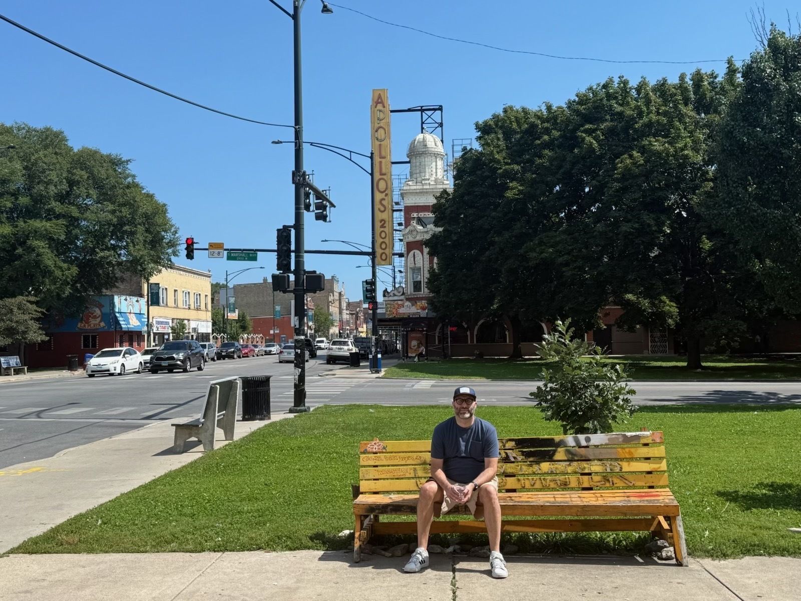 Man wearing a cap, glasses, navy shirt, and shorts sitting on a yellow graffiti-covered bench in a green park near a street with cars and the Apollo 2000 theater sign in the background.