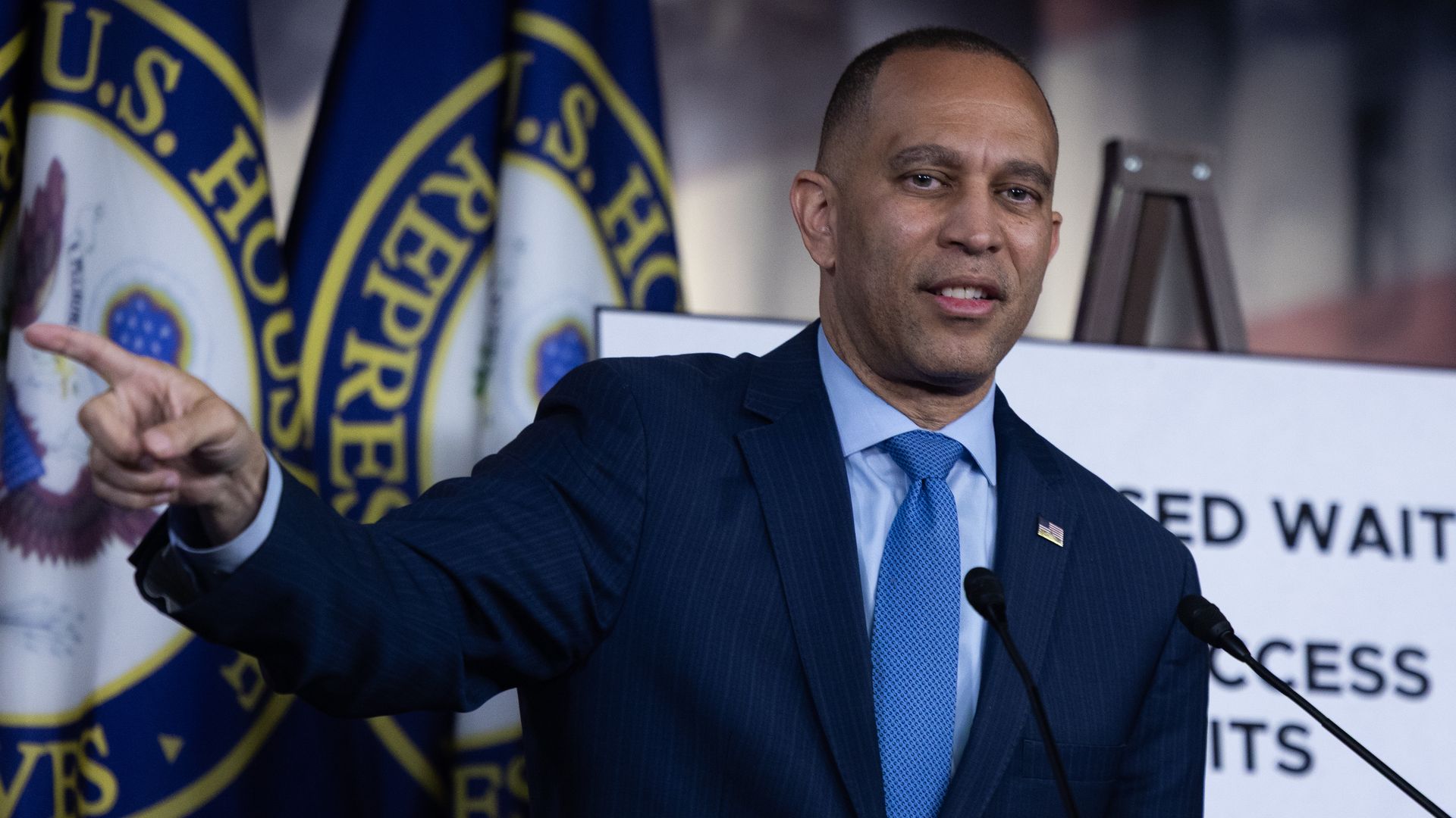 Hakeem Jeffries, wearing a blue suit and speaking into microphones in front of blue flags and a white poster.