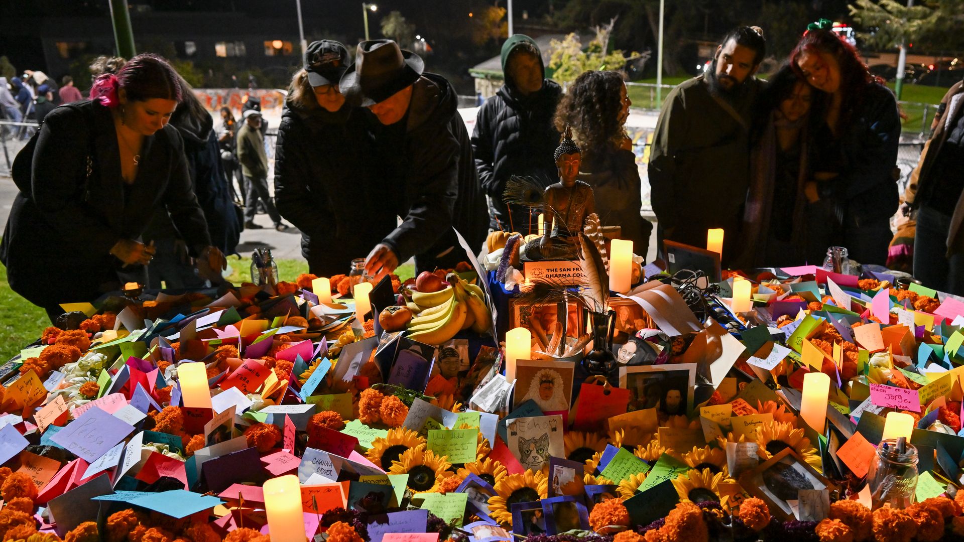 People gathered at night around a colorful altar with marigolds, sunflowers, candles, photos, and notes in tribute, for a memorial or Day of the Dead celebration.