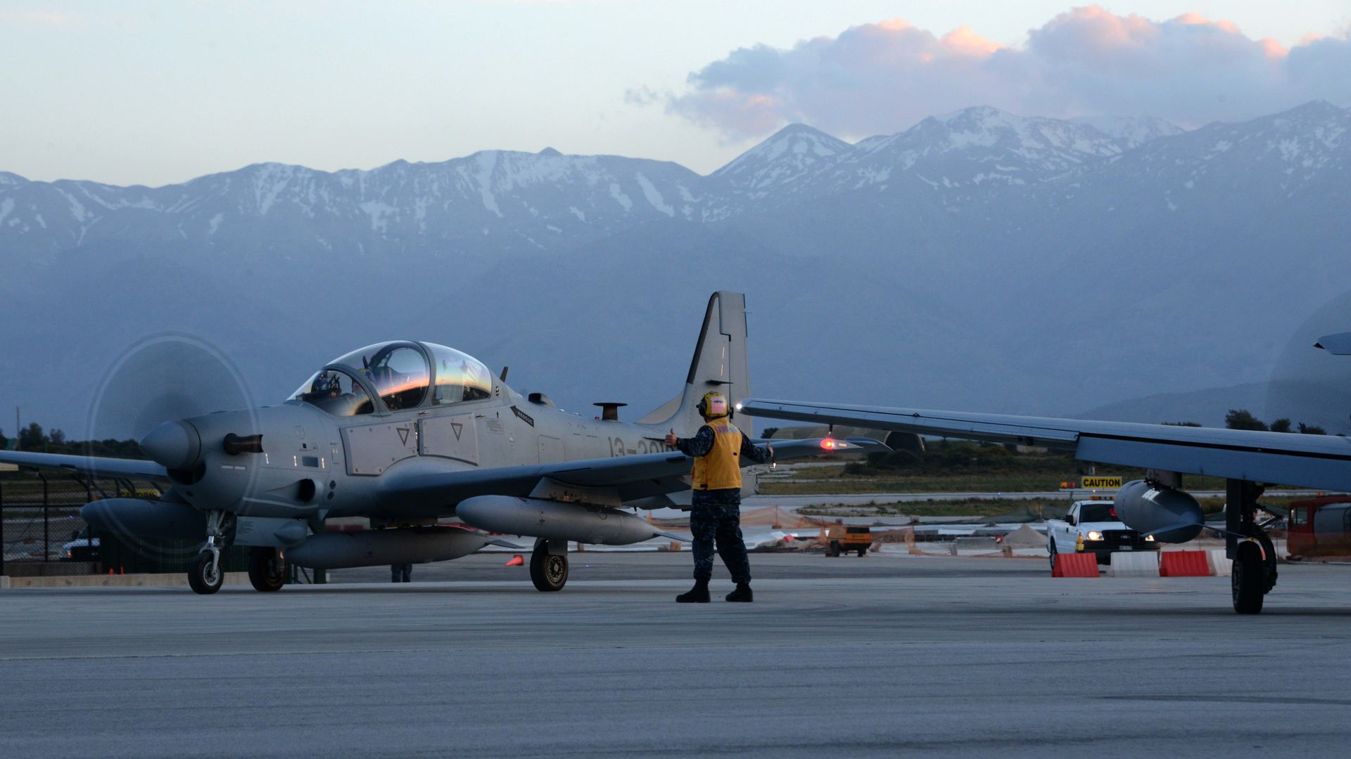 Gray military aircraft on runway at dusk with mountains in background and personnel in yellow vest directing the plane