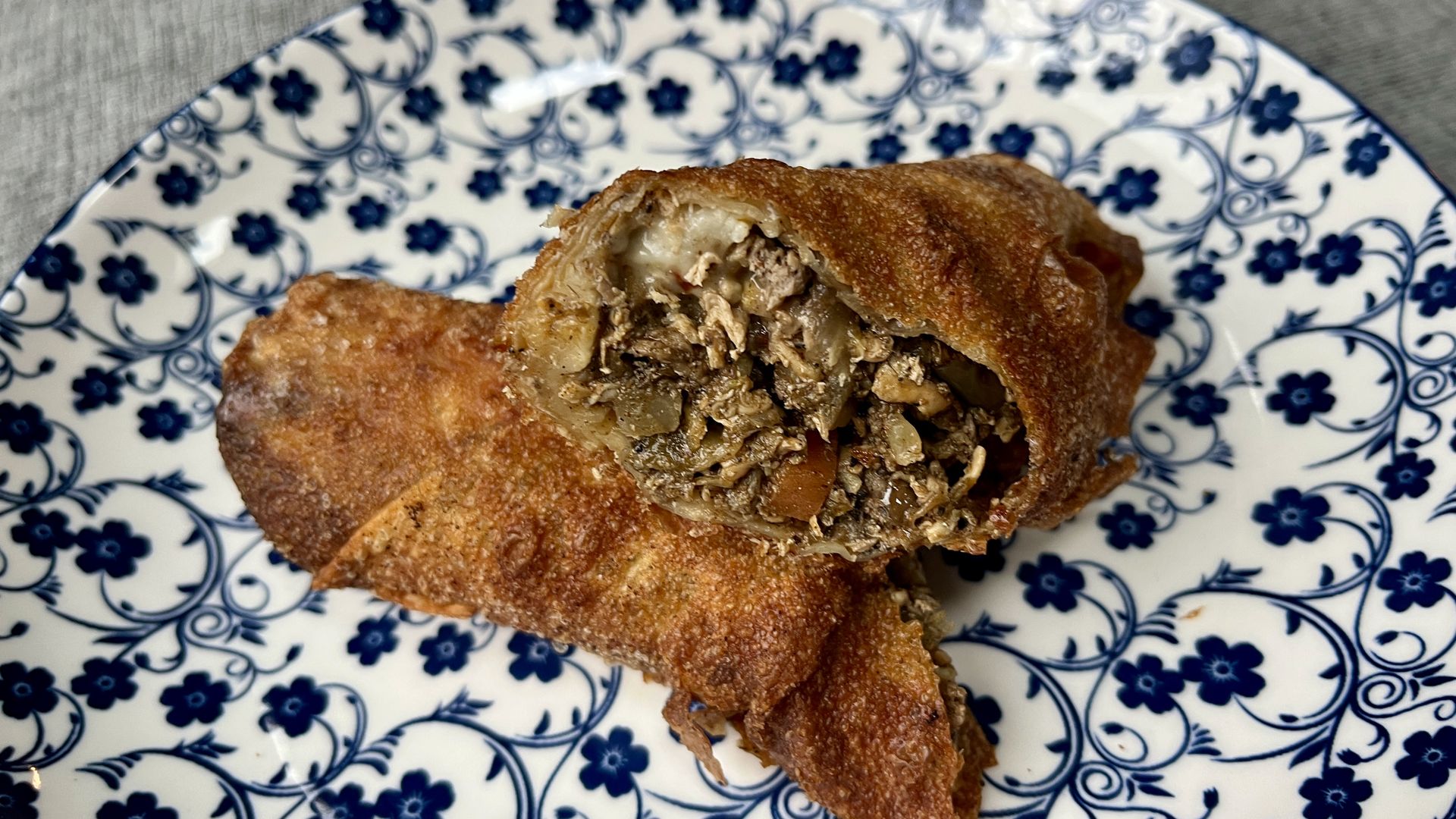 Two crispy golden-brown egg rolls on a white plate with blue floral patterns, one cut open to show a filling of vegetables and shredded meat.