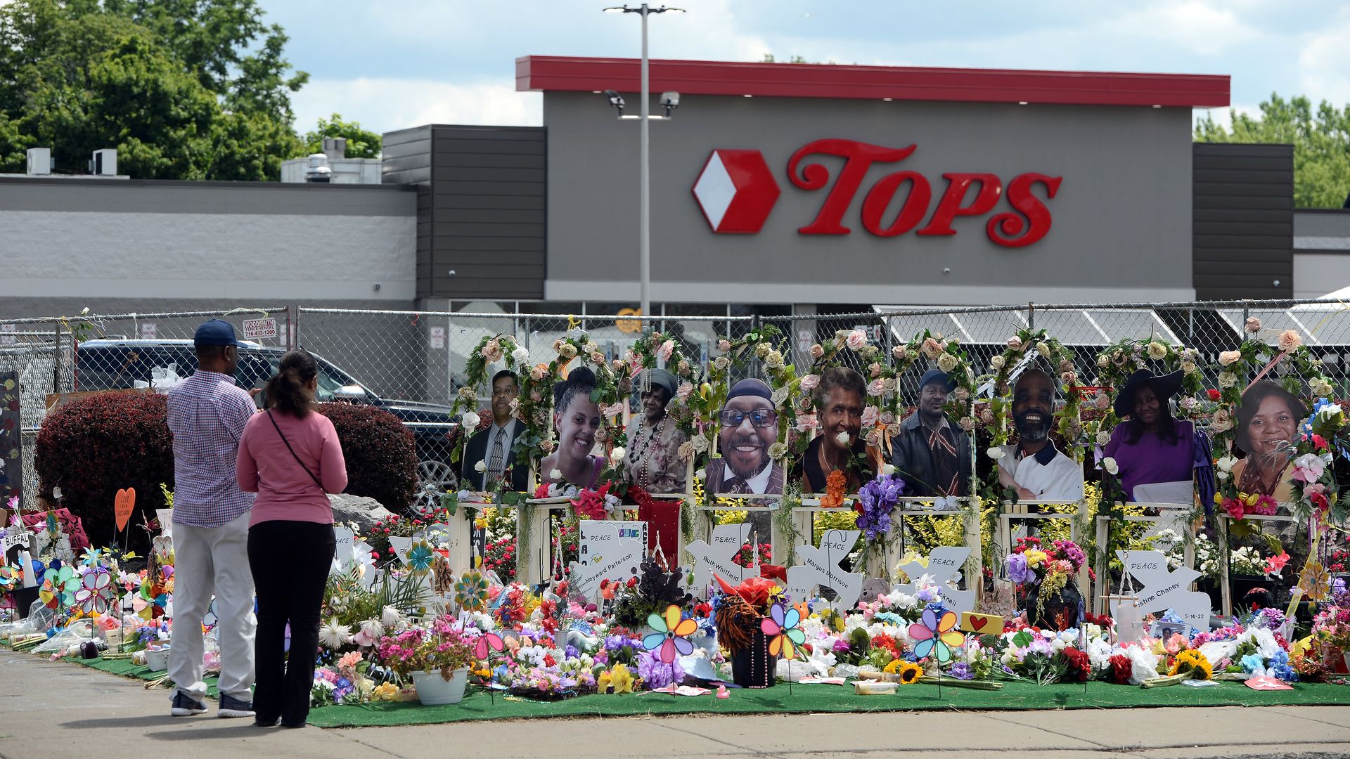 Photo of flowers, pictures and signs positioned in front of the Tops grocery store as two people look at the memorial site