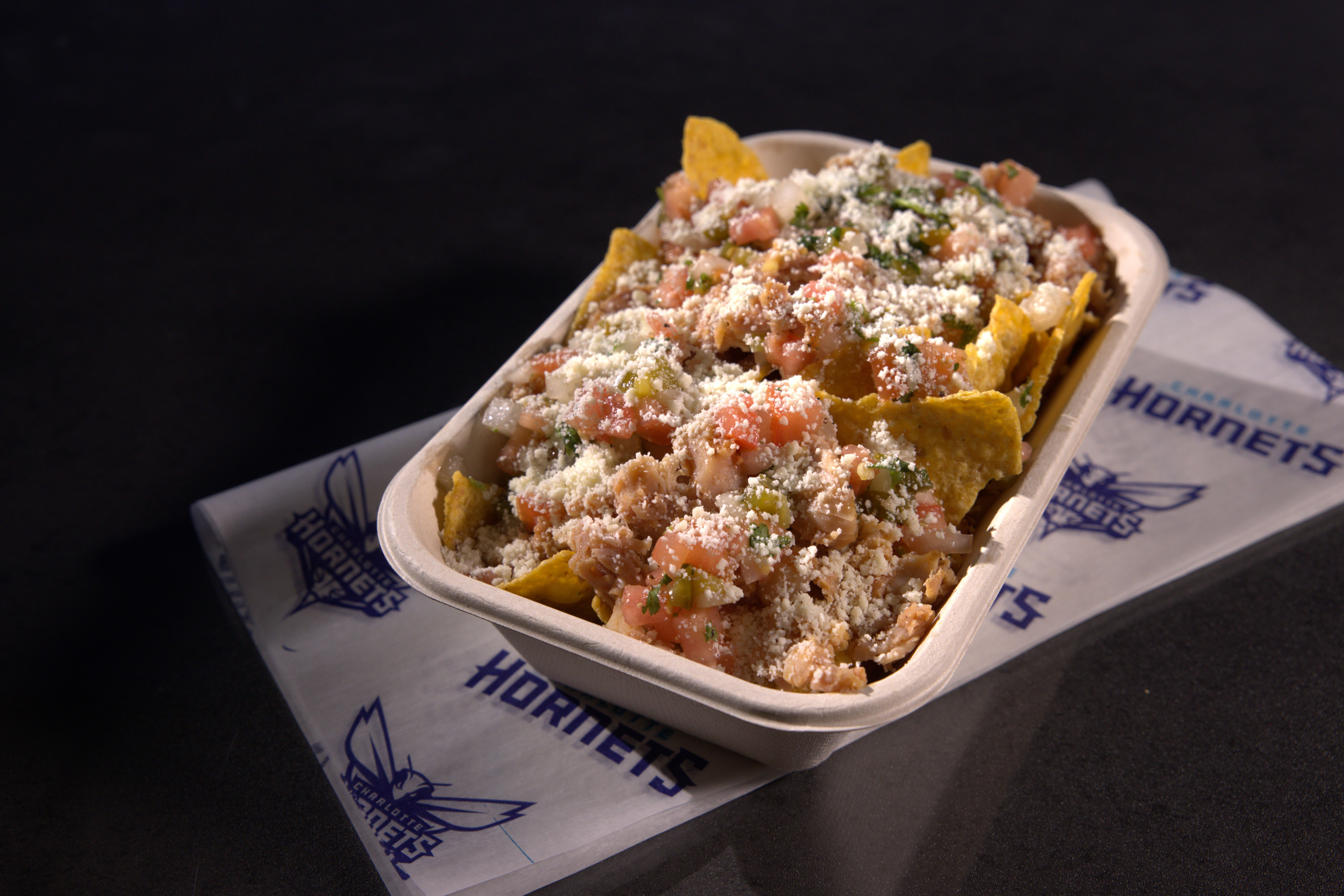 A tray of nachos topped with diced tomatoes, onions, cilantro, shredded chicken, and white crumbled cheese on a Charlotte Hornets branded napkin against a dark background.