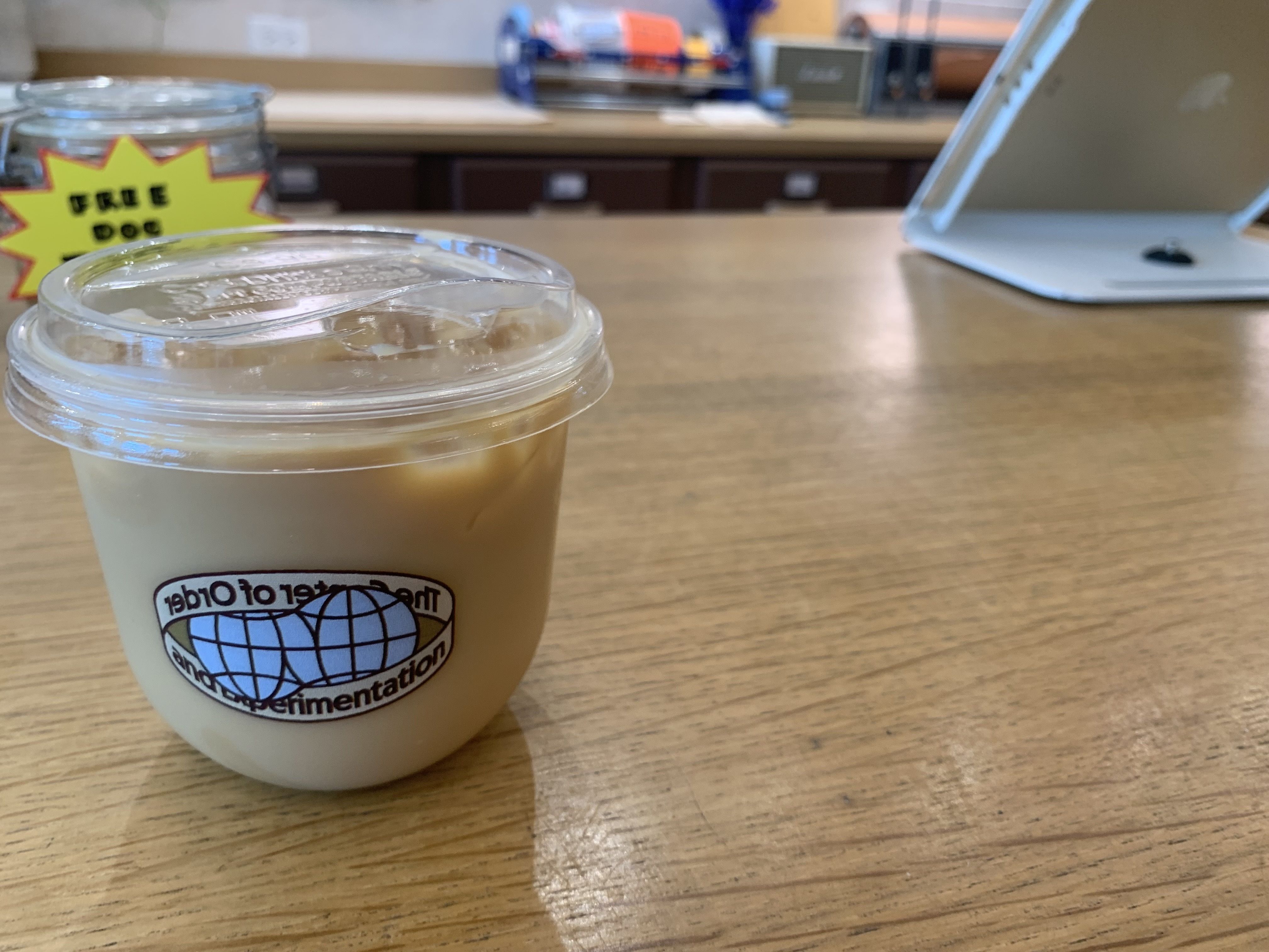 Small clear plastic cup with iced coffee on wooden counter, labeled with a blue globe and text about order and experimentation, with a blurred background of a jar and white stand.