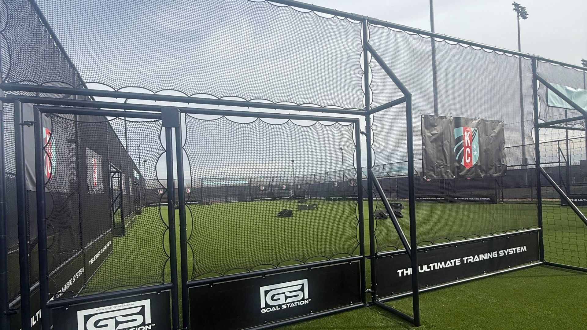 Outdoor soccer training area with artificial green turf enclosed by black netting and frames, featuring Goal Station branding and a black banner with a red and blue KC shield logo.