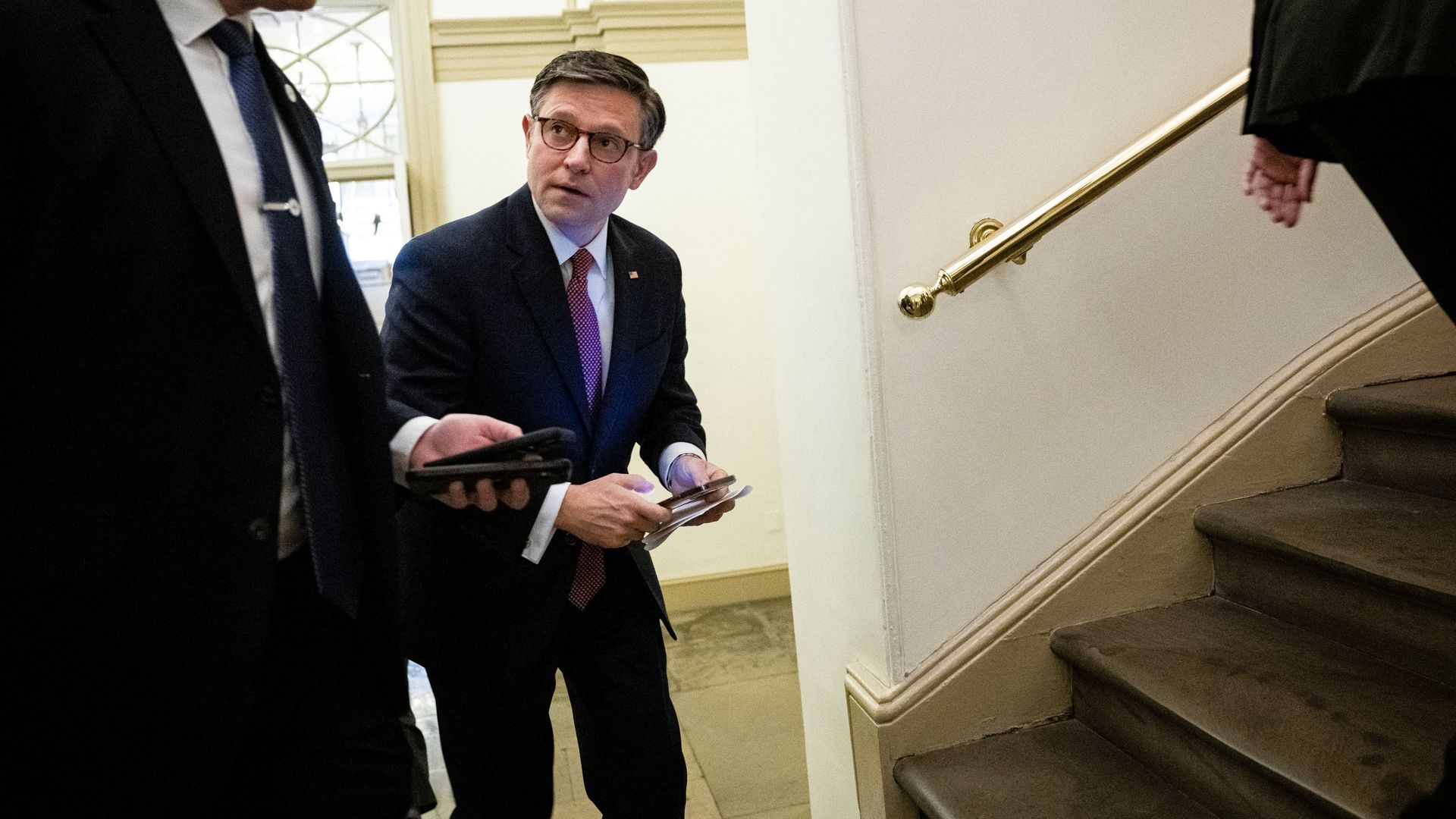 House Speaker Mike Johnson, wearing  a blue suit and glasses, walking towards stairs in the Capitol.