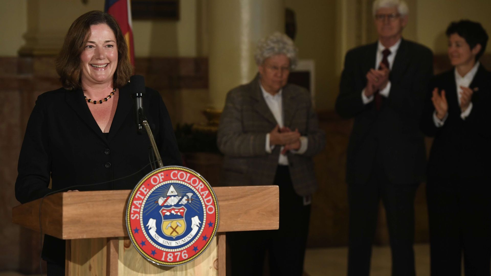 Smiling woman in a black blazer speaks at a podium adorned with the Colorado seal; a small crowd claps in a formal indoor setting with a flag in the background.
