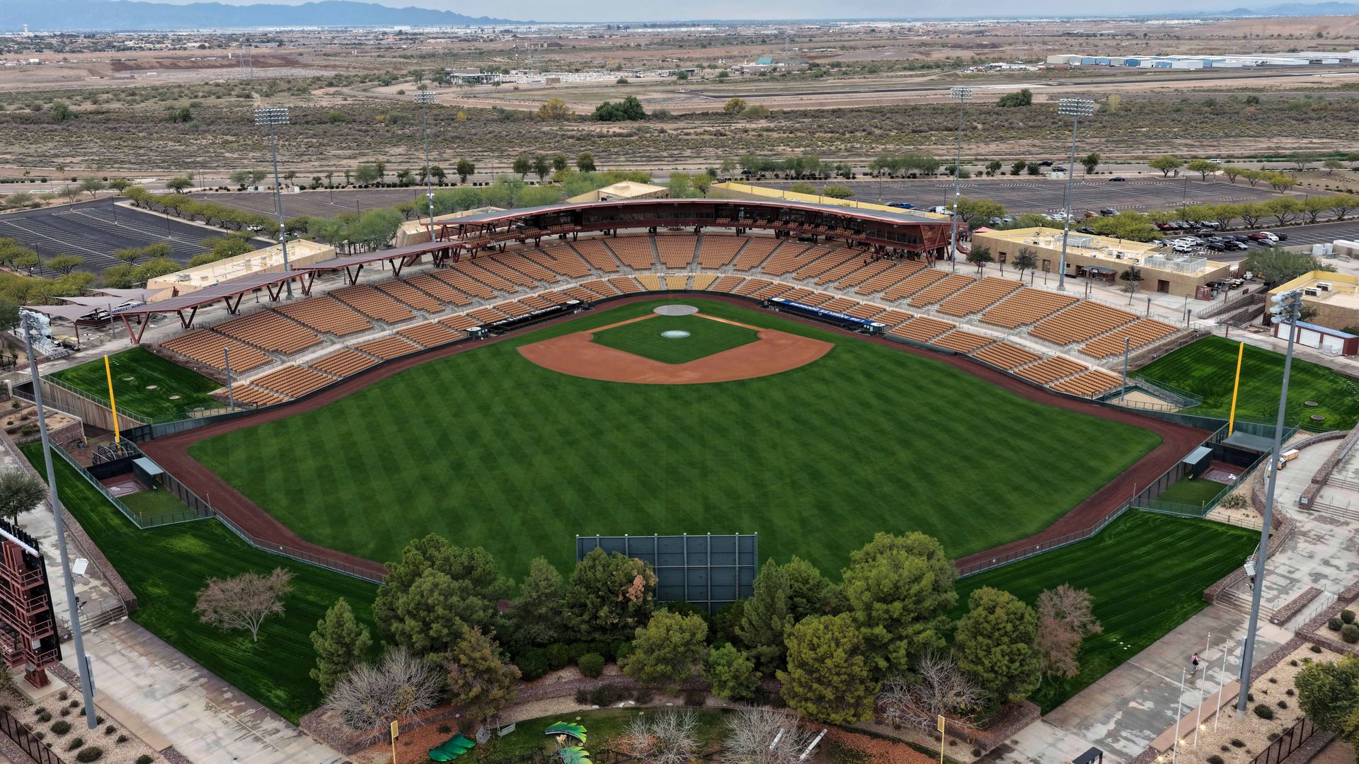 Aerial view of an empty baseball stadium with lush green field and brown dirt infield, tan seats, surrounded by parking lots and desert landscape under a cloudy sky.