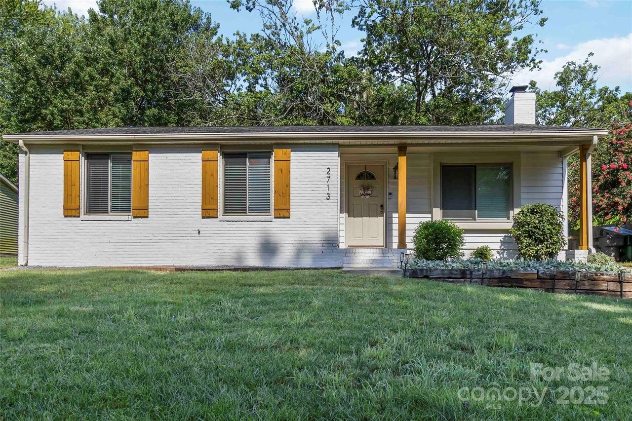 Single-story white brick house with yellow wooden shutters, a small porch with wooden columns, and green lawn surrounded by trees under a blue sky.
